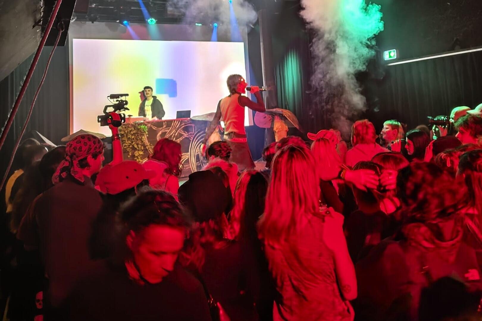 Ein Künstler singt auf der Bühne mit einem DJ und einem Kameramann im Hintergrund, während eine lebhafte Menge zusieht. Bunte Bühnenlichter und Raucheffekte verstärken die lebhafte Konzertatmosphäre.