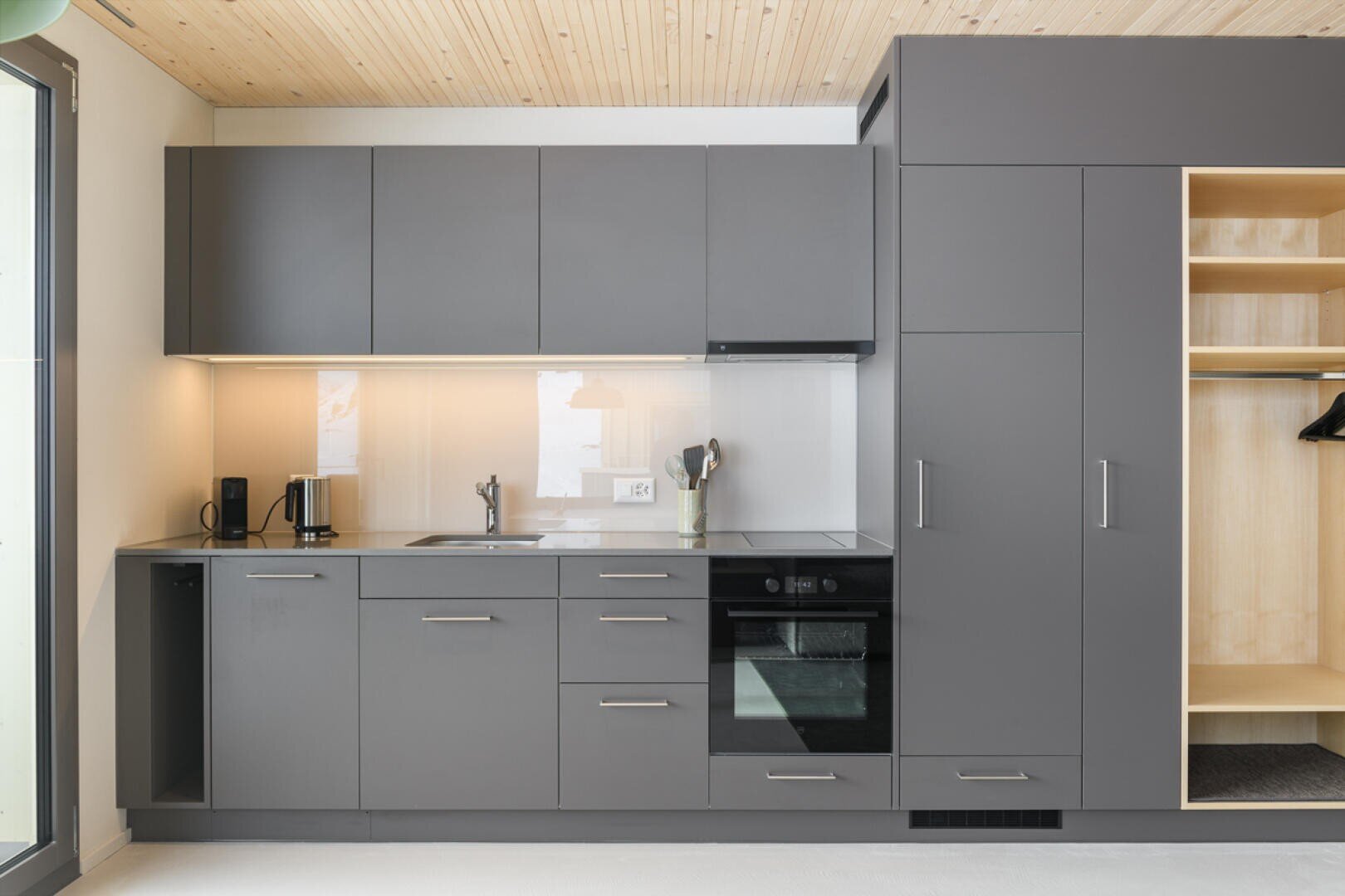 Moderne Küche mit mattgrauen Schränken, Einbauherd, offenen Regalen, Spüle, Kaffeemaschine und minimalem Dekor. Eine helle Holzdecke und ein teilweise offener Kleiderschrank sind ebenfalls zu sehen. Reichlich natürliches Licht fällt von links ein.