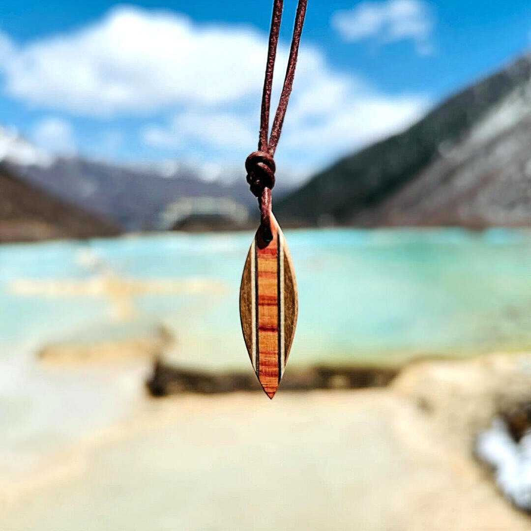 An einer braunen Kordel hängt ein kleiner hölzerner Surfbrett-Anhänger, im Hintergrund ein türkisfarbener See und verschwommene Berge unter einem teilweise bewölkten Himmel.