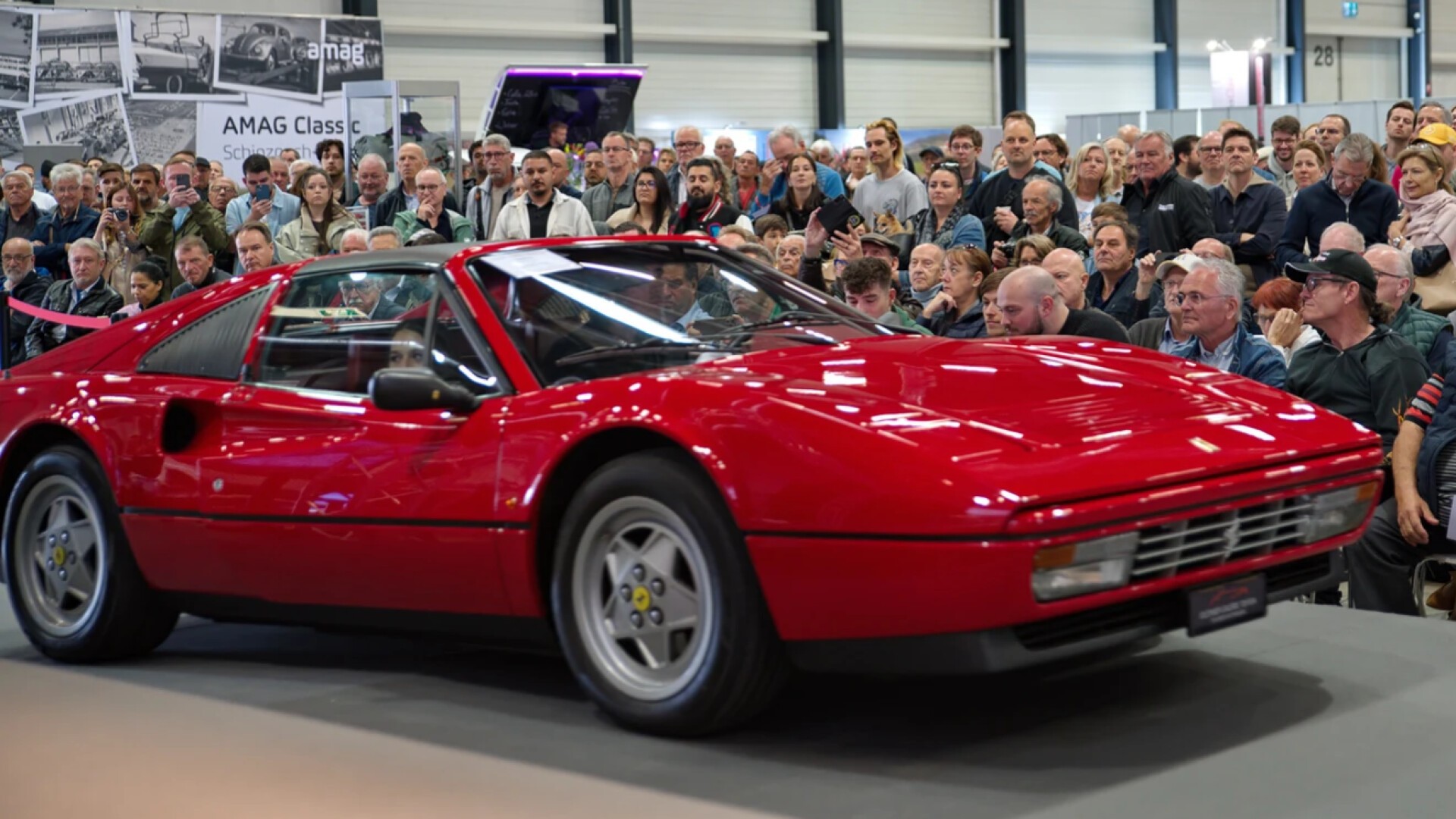 Ein klassischer roter Ferrari-Sportwagen wird in einem Innenbereich ausgestellt, umgeben von einer großen Menschenmenge, die das Auto bei einer Automobilveranstaltung oder -ausstellung aufmerksam beobachtet und fotografiert.