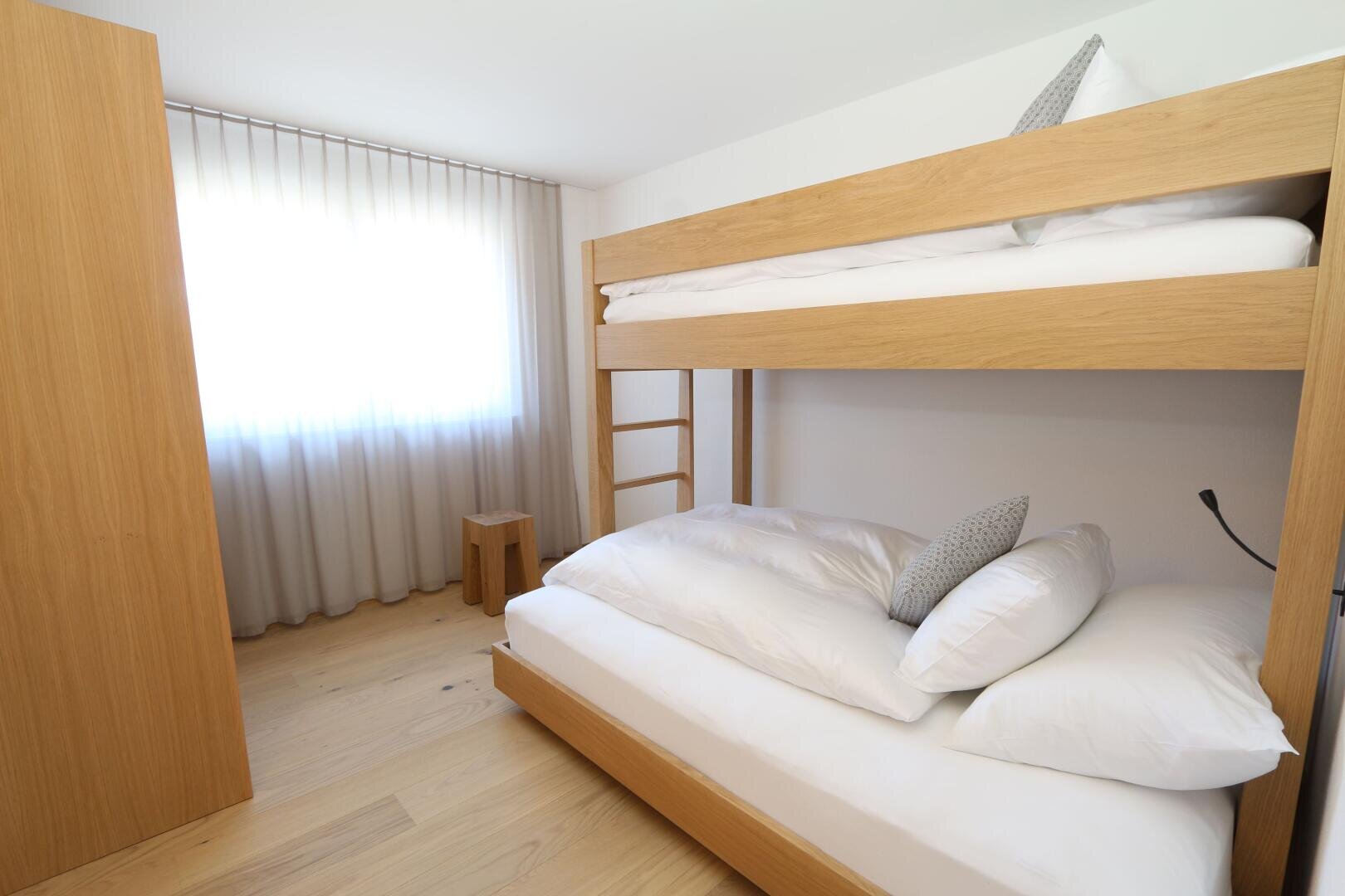 Ein helles, minimalistisches Schlafzimmer mit einem Etagenbett aus Holz, jedes Bett ordentlich mit weißer Bettwäsche und einem grauen Kissen bezogen. Das Licht fällt durch die durchsichtigen Vorhänge, die ein großes Fenster verdecken. Ein Holzhocker steht neben dem Fenster.