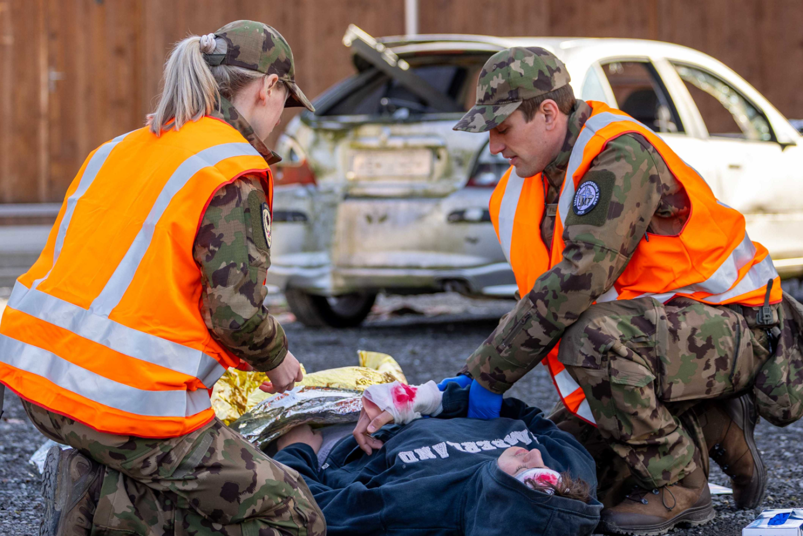 Zwei Personen in Tarnuniformen und orangefarbenen Sicherheitswesten kümmern sich um eine verletzte Person, die mit bandagierten Armen am Boden liegt. Im Hintergrund ist ein beschädigtes Auto zu sehen. Es scheint sich um einen Notfall oder Erste-Hilfe-Maßnahmen zu handeln.
