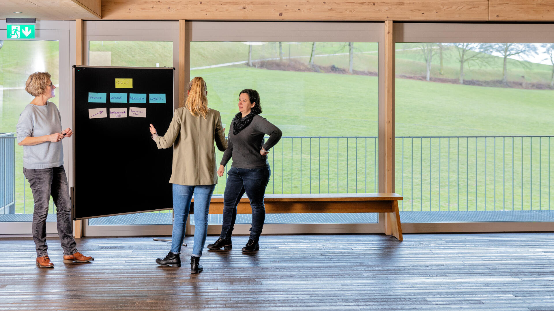 Drei Personen stehen in einem modernen, hellen Raum mit großen Fenstern und Ausblick auf die Landschaft und diskutieren über Notizen an einer Tafel. Einer schreibt, während die anderen beobachten und sich unterhalten.