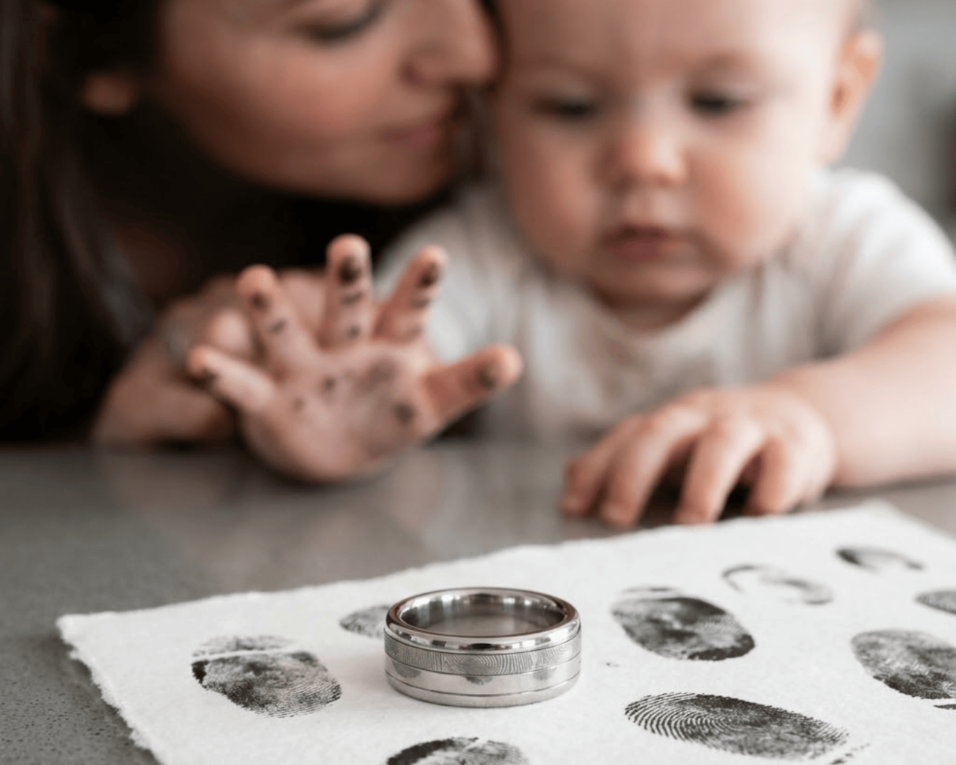 Ein Baby mit schmutzigen Fingern greift nach zwei Ringen auf einem Tuch mit Fingerabdrücken, während sich eine erwachsene Frau ganz nah heranbeugt, die im Hintergrund verschwommen zu sehen ist.