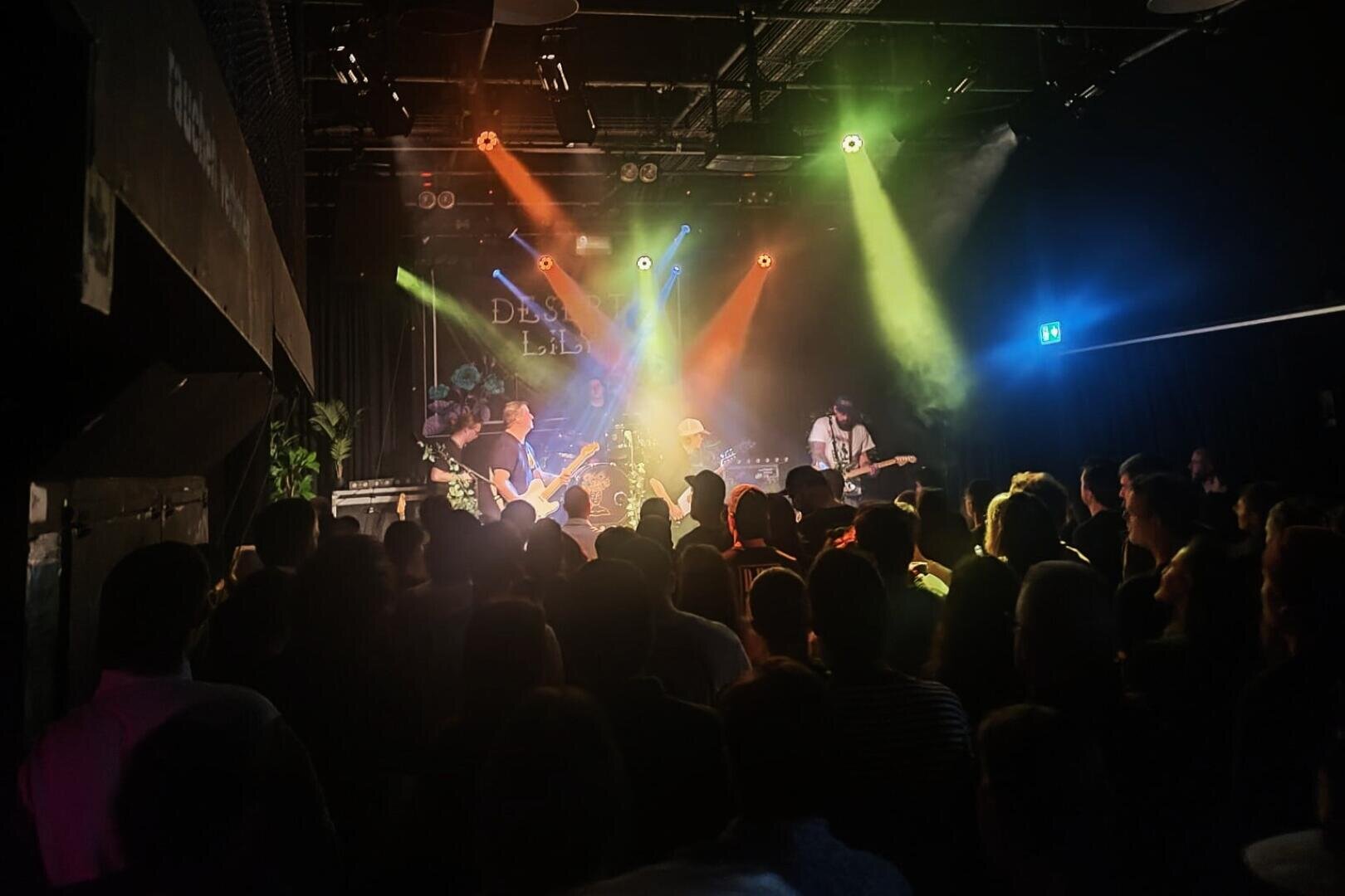 Eine Band spielt auf der Bühne unter bunten Lichtern in einer dunklen Halle, während eine Menschenmenge zusieht. Der Bühnenhintergrund zeigt schwachen Text und Grafiken.