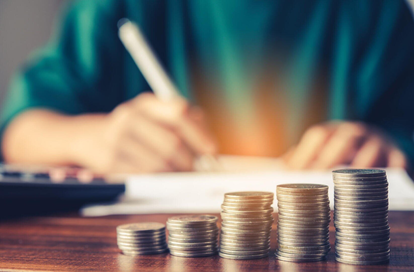 Stapel von Münzen in ansteigender Höhe auf einem Tisch mit einer unscharfen Person im Hintergrund, die auf Papier schreibt, was auf Finanzplanung oder Geldsparen hindeutet.