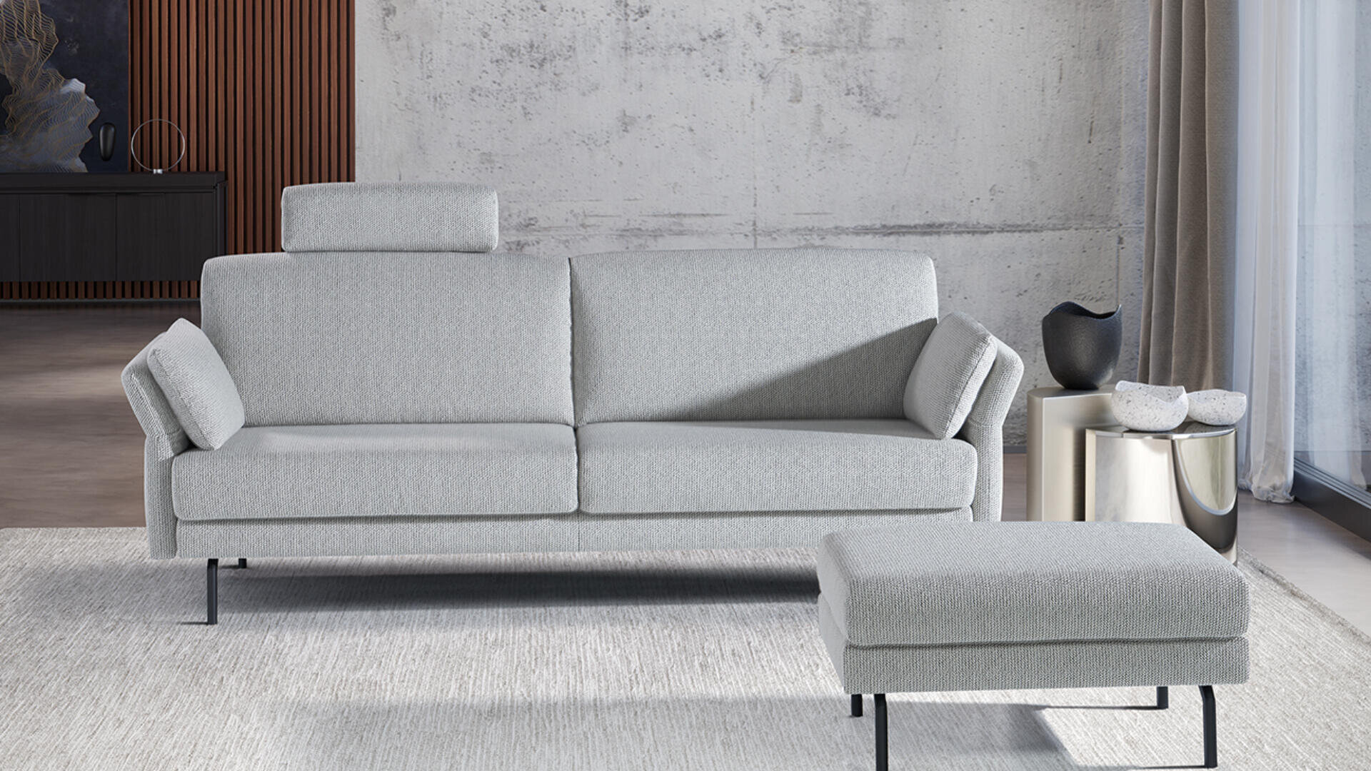 Ein modernes Wohnzimmer mit einem hellgrauen Sofa mit verstellbaren Armlehnen und einer passenden Ottomane auf einem hellen Teppich. Der Hintergrund besteht aus einer Betonwand, einem Vorhang und dekorativen Möbelstücken.