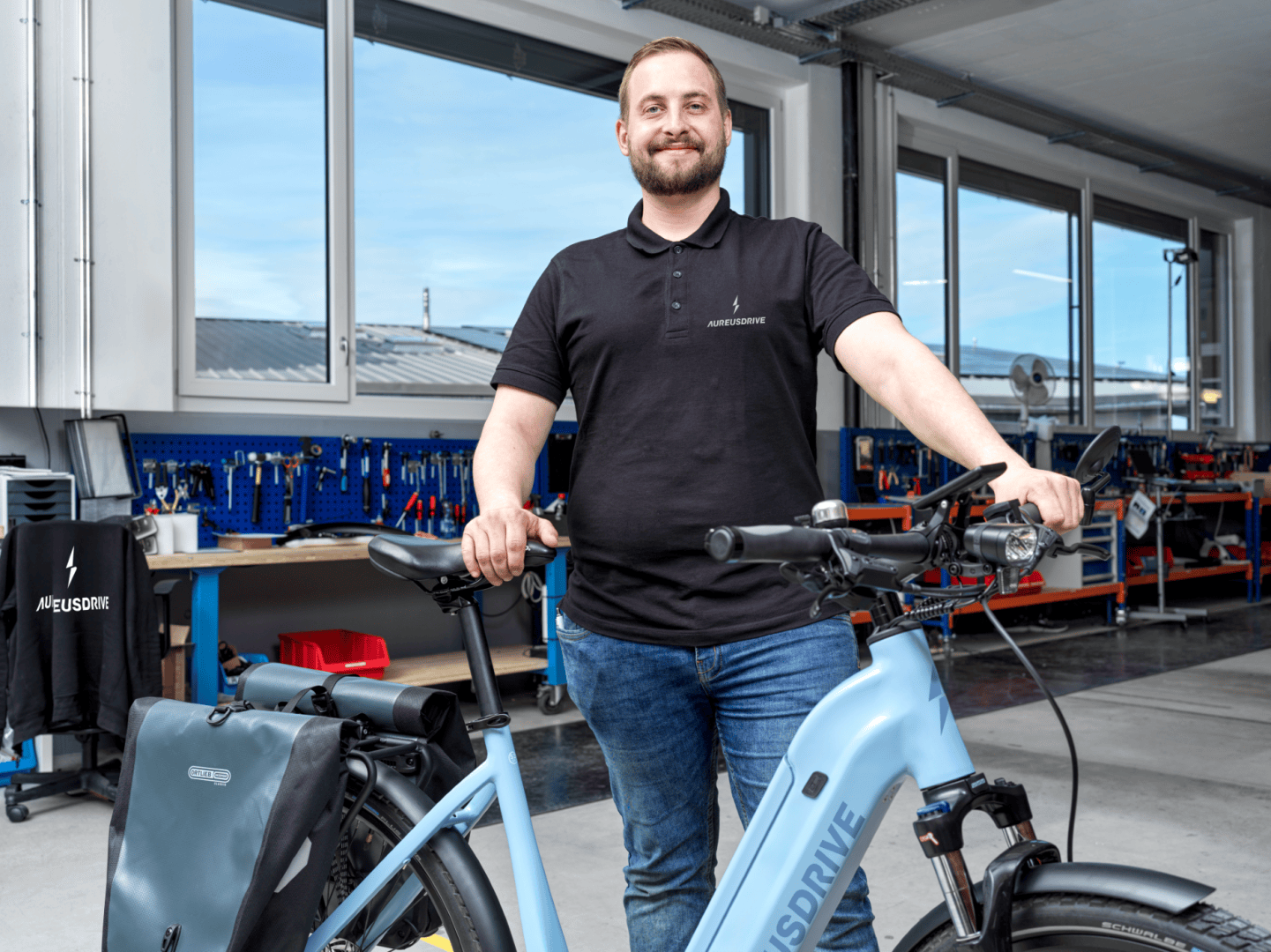 Ein lächelnder Mann in einem schwarzen Poloshirt steht in einem Raum und hält den Lenker eines hellblauen Elektrofahrrads. Der Raum hat große Fenster und Werkbänke mit Werkzeugen und Geräten im Hintergrund.