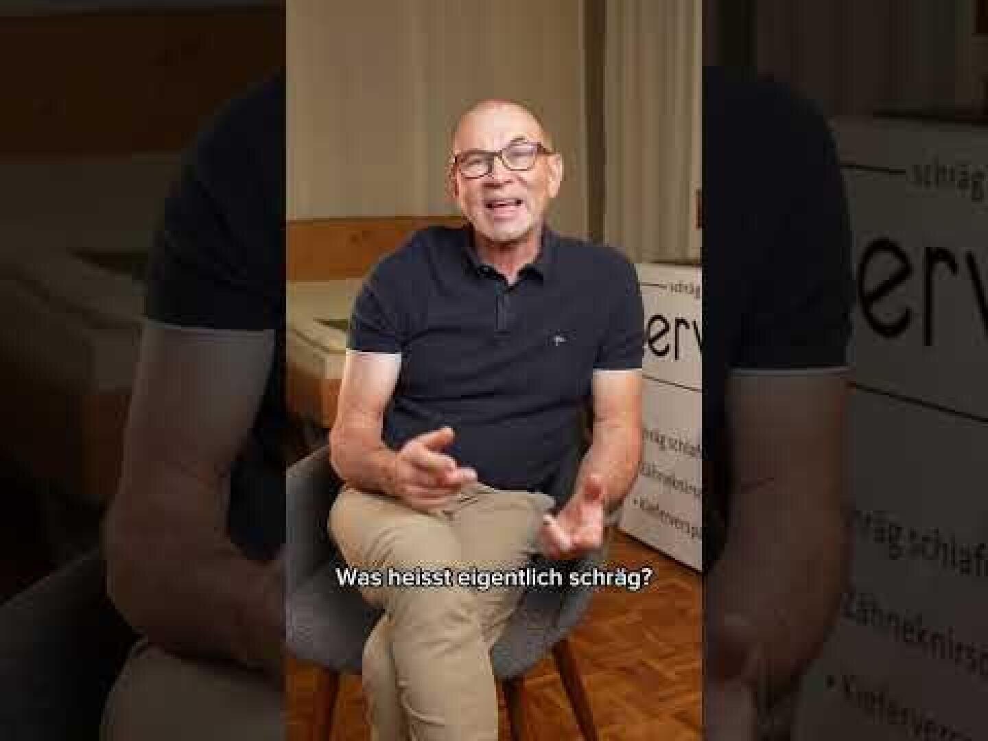 Ein lächelnder, glatzköpfiger Mann mit Brille, dunklem Polohemd und beiger Hose sitzt auf einem Stuhl in einem Innenraum. Er gestikuliert mit seinen Händen, während er spricht. Die deutschen Untertitel am unteren Bildrand lauten: Was heißt eigentlich schräg?.