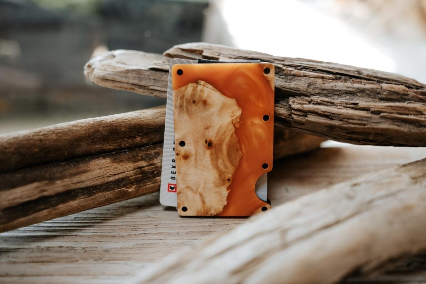 Ein minimalistisches Portemonnaie mit einem einzigartigen orange-beigen Holzmaserungsdesign ruht aufrecht zwischen glatten, verwitterten Treibholzstücken auf einer Holzoberfläche.