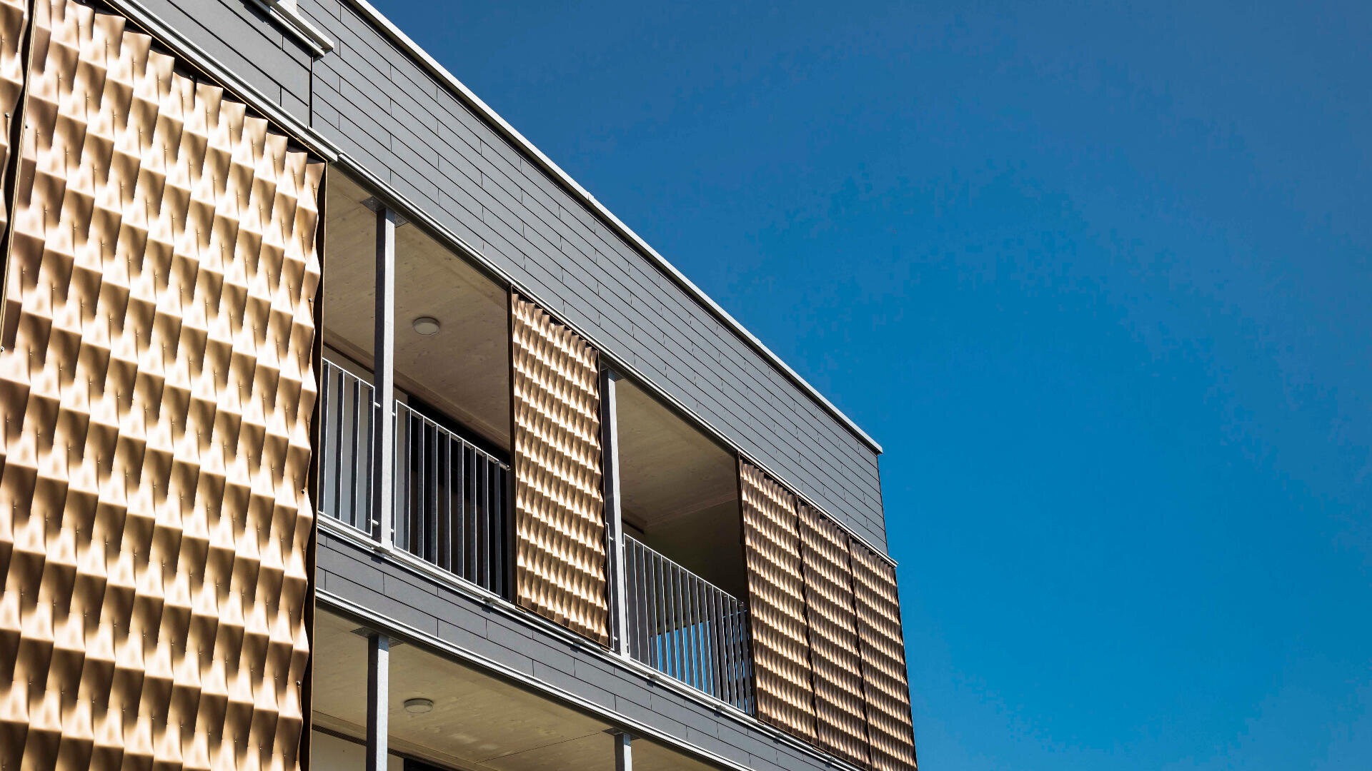 Moderne Gebäudefassade mit strukturierten, gewellten Paneelen und Metallgeländern vor einem klaren blauen Himmel. Das Gebäude verfügt über Balkone und eine zeitgenössische architektonische Gestaltung.