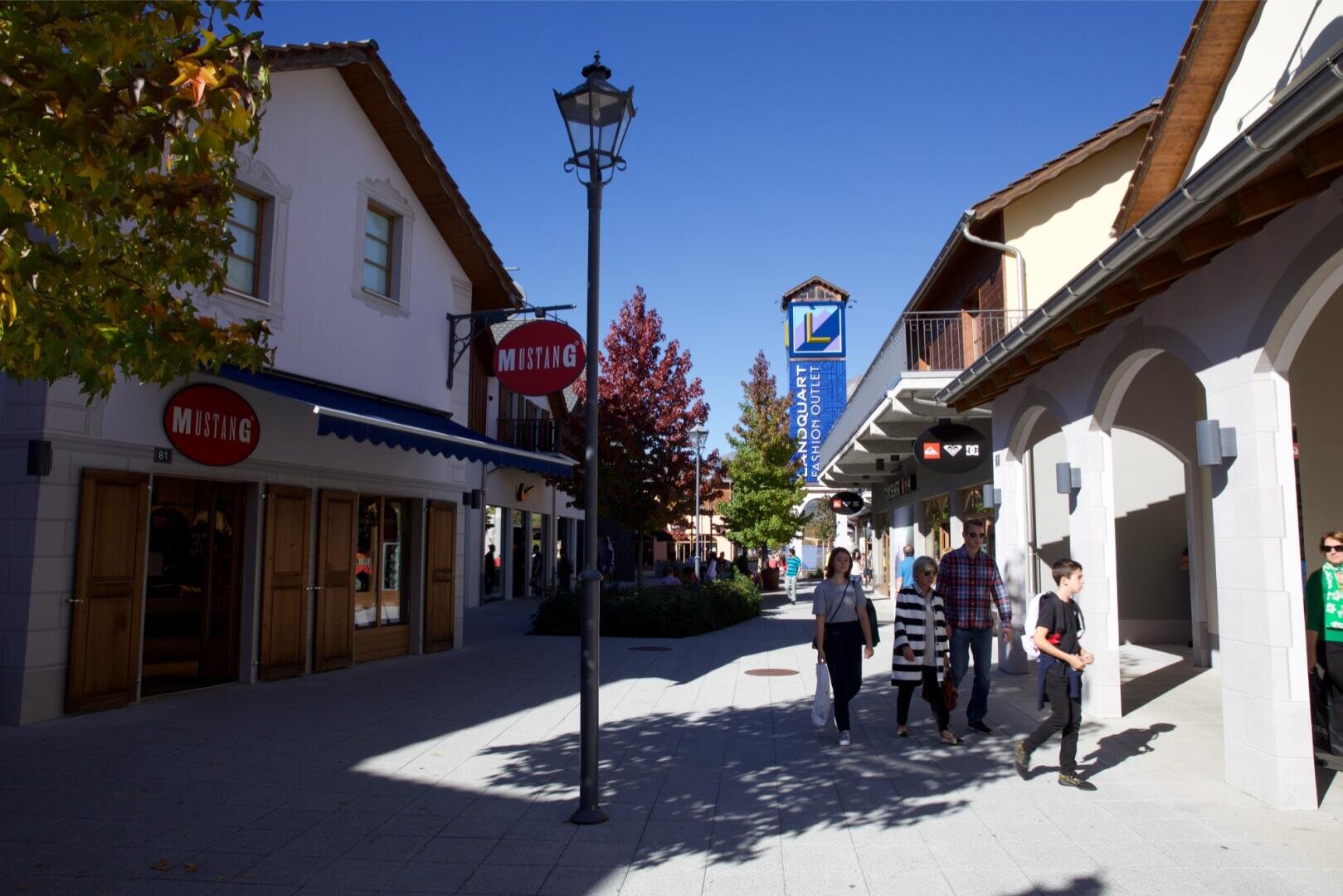Menschen gehen eine sonnige Fußgängerzone entlang, die von Geschäften und Bäumen gesäumt ist. Die Schaufenster sind mit Schildern versehen, darunter eines für MUSTANG. Schatten und Herbstlaub schaffen eine lebendige, entspannte Atmosphäre.