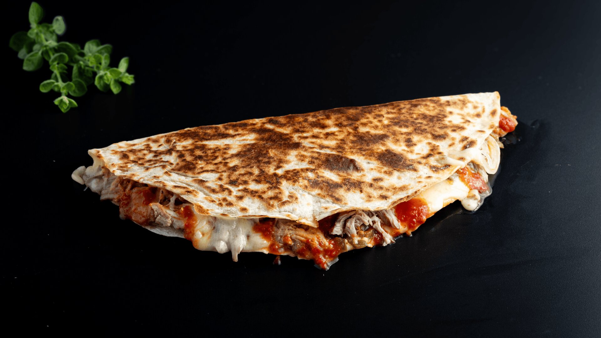 Eine gegrillte Quesadilla, gefüllt mit geschmolzenem Käse, geschnetzeltem Fleisch und Tomatensoße, liegt auf einer schwarzen Unterlage, daneben liegt ein Zweig frischer Kräuter.