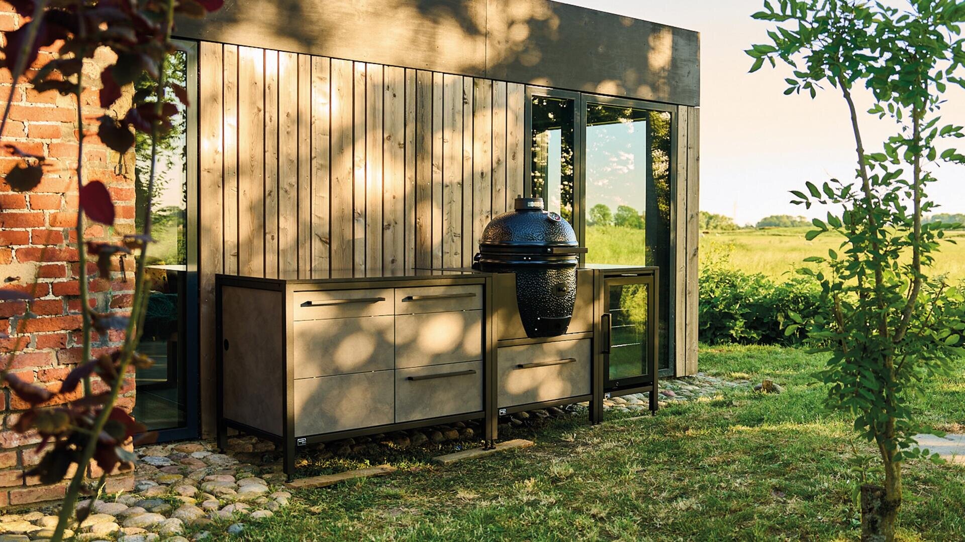 Eine moderne Außenküche mit einem Grill im Kamado-Stil steht neben einem Holzhaus mit großen Glastüren, umgeben von grünem Gras und Gartenpflanzen im warmen Sonnenlicht.