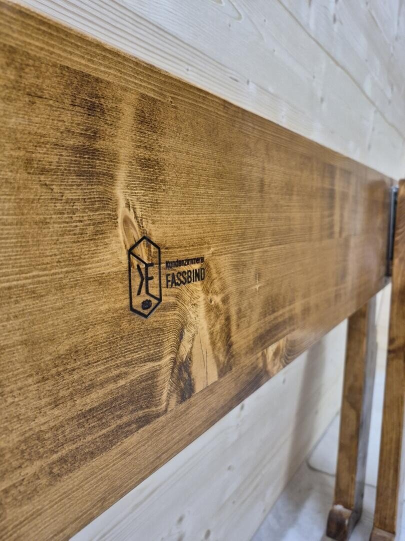 Nahaufnahme einer Holzbank, auf deren Seite das Logo und der Text Fassbind eingraviert sind, vor einer hellen holzgetäfelten Wand. Die Bank hat eine glatte, polierte Oberfläche, die die Holzmaserung hervorhebt.