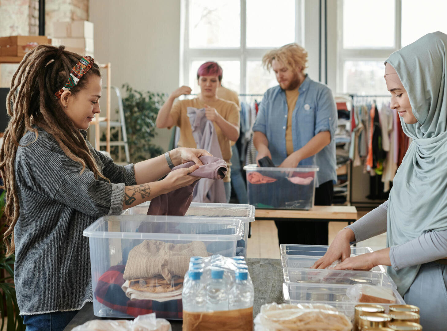 Vier Personen sortieren und falten Kleidung in durchsichtigen Plastikbehältern in einem hellen Raum. Zwei Frauen arbeiten im Vordergrund, während zwei andere im Hintergrund die Kleidung sortieren. Auf dem Tisch stehen Wasser in Flaschen und abgepackte Lebensmittel.