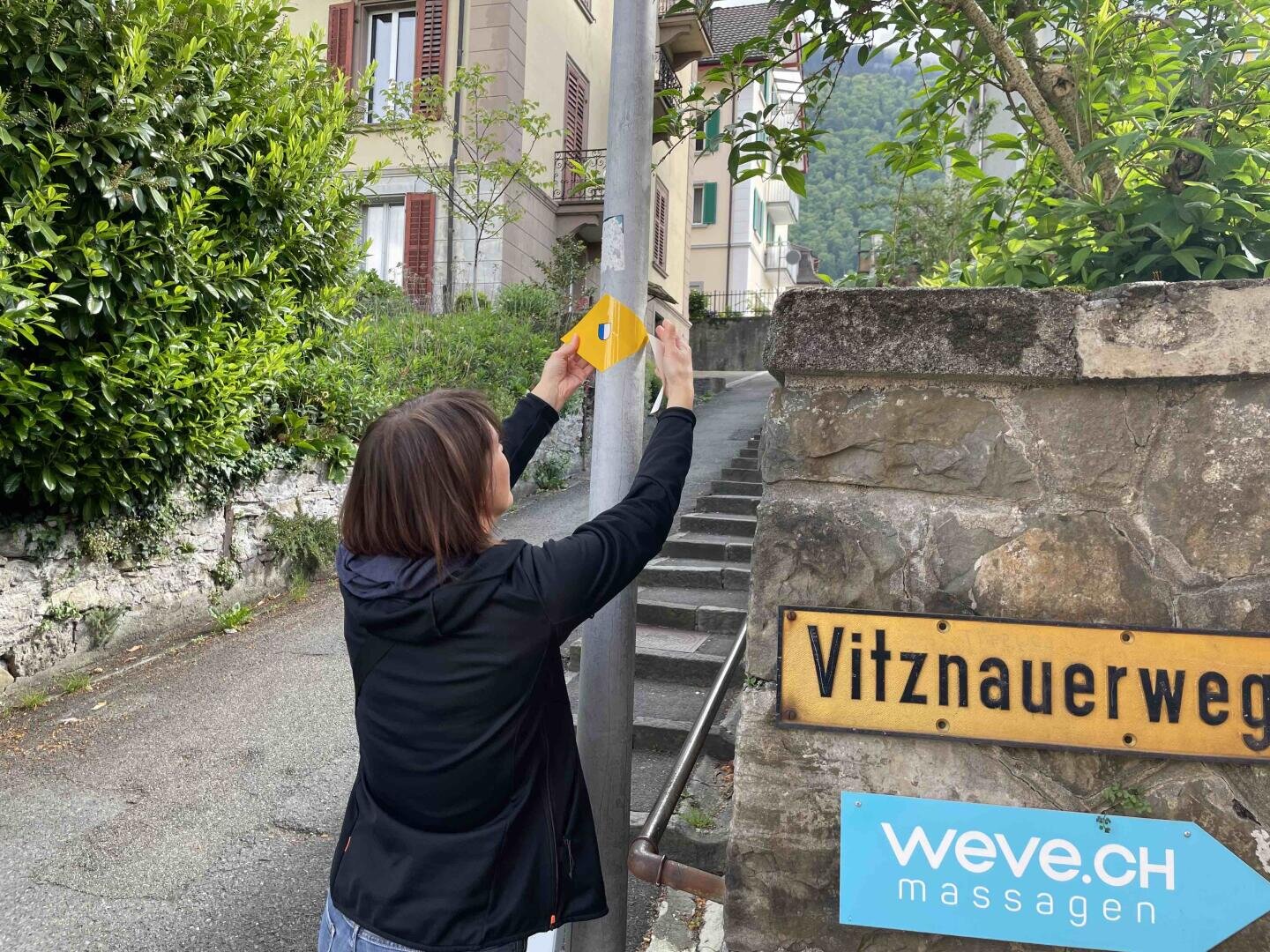 Eine Frau befestigt eine gelbe Wegmarkierung an einem Pfahl in der Nähe von Steinstufen an einer Straßenecke. Auf den Schildern an der Wand steht Vitznauerweg und weve.ch massagen. Im Hintergrund sind Wohnhäuser und Grünanlagen zu sehen.