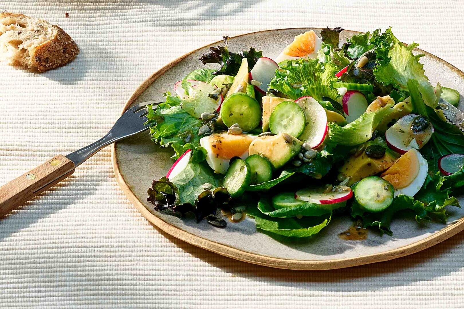 Ein Teller mit frischem Salat mit Blattgemüse, geschnittenen Gurken, Radieschen, hart gekochten Eiern, Kürbiskernen und einem leichten Dressing. Eine Gabel und ein Stück Brot liegen daneben auf einer strukturierten, hellen Tischdecke.
