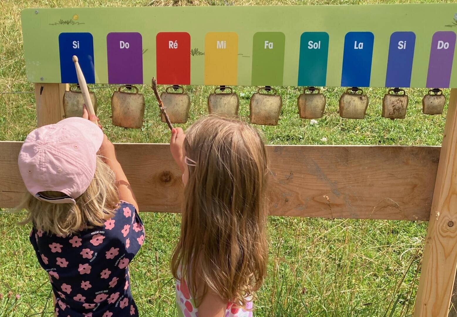 Zwei junge Mädchen, eines mit einem rosa Hut, spielen auf einer Wiese mit Stöcken ein Musikinstrument mit beschrifteten bunten Stäben (Si, Do, Ré, Mi, Fa, Sol, La, Si, Do) und Kuhglocken.