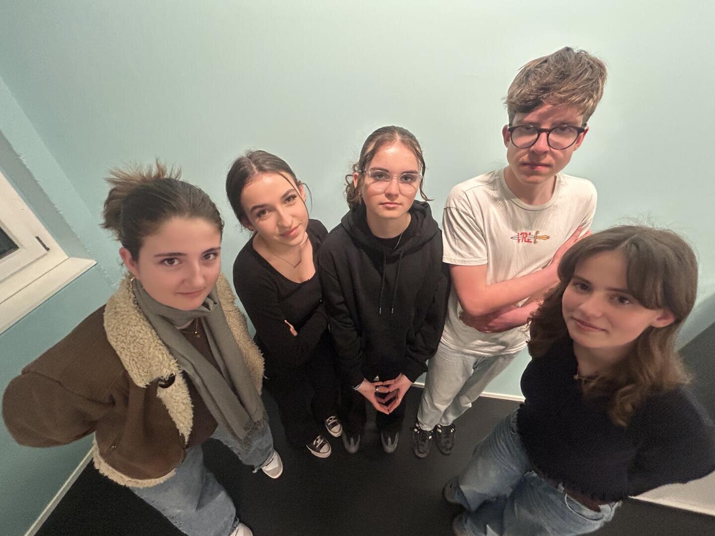 Fünf junge Menschen stehen in einem Innenraum eng beieinander, von oben betrachtet. Vier tragen lässige Oberteile und Jeans, einer trägt eine Brille und ein weißes T-Shirt. Die Gruppe steht vor einer hellblauen Wand und blickt in Richtung der Kamera.