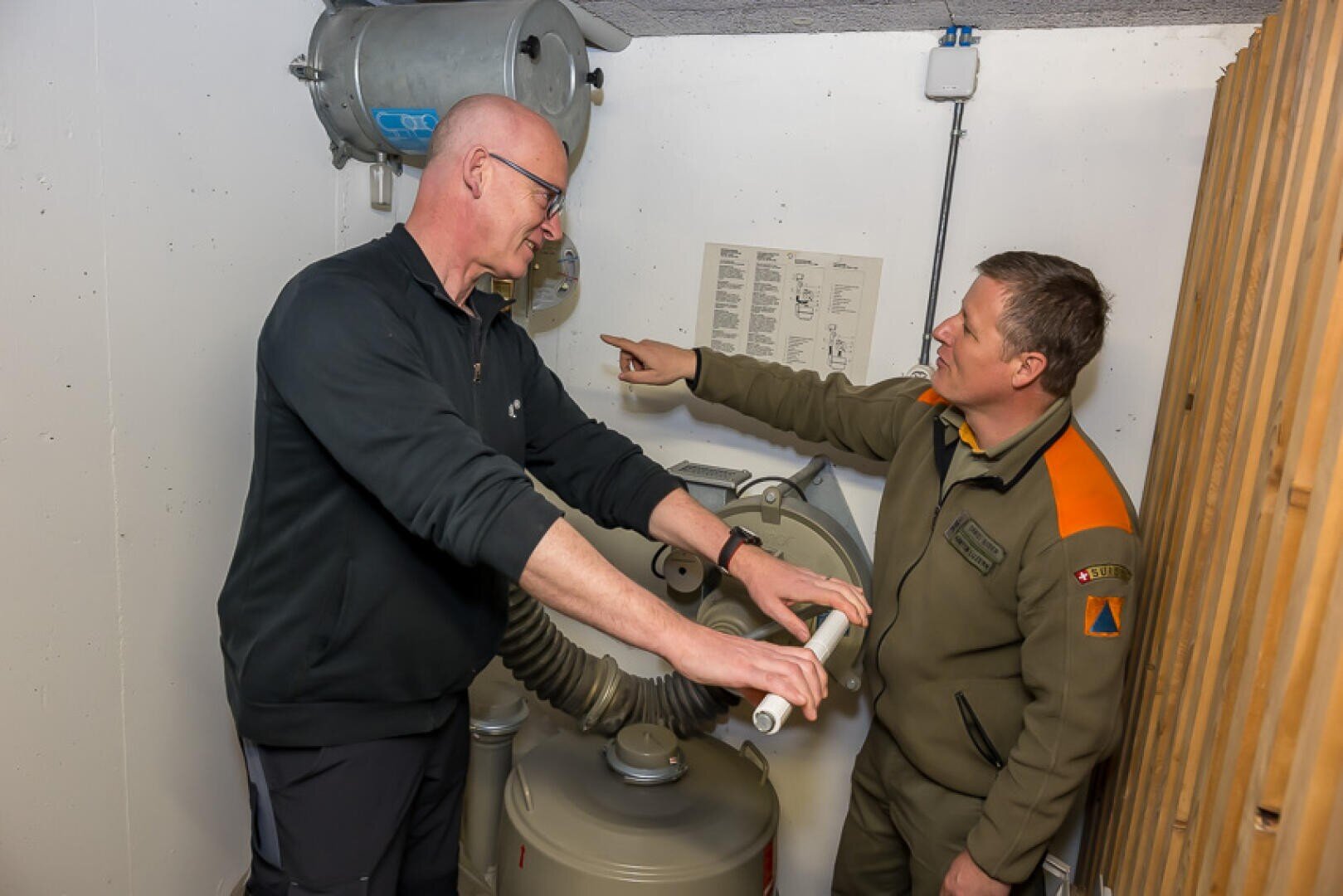 Zwei Männer stehen in einem kleinen Raum mit Maschinen; ein Mann bedient eine große Handkurbel, während der andere, der in Uniform gekleidet ist, auf Anweisungen an der Wand zeigt und scheinbar eine Anleitung gibt.