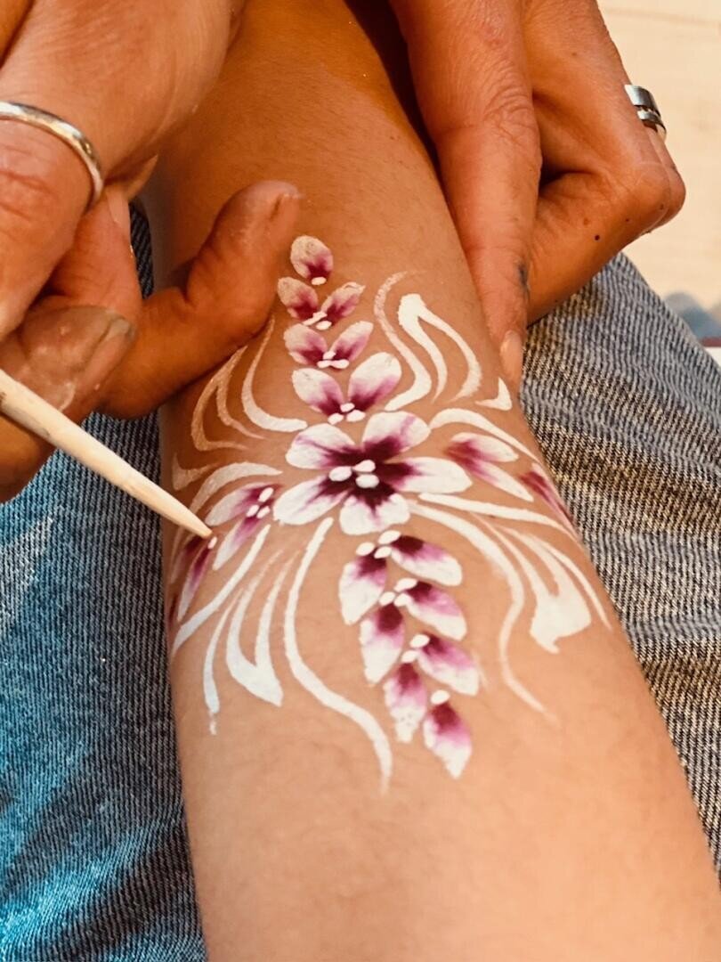 Eine Hand hält einen Stock und malt mit weißen und violetten Farben ein florales Muster auf den Unterarm einer Person, wobei komplizierte Blütenblätter und Muster entstehen. Die Person, die bemalt wird, trägt Denim-Jeans.