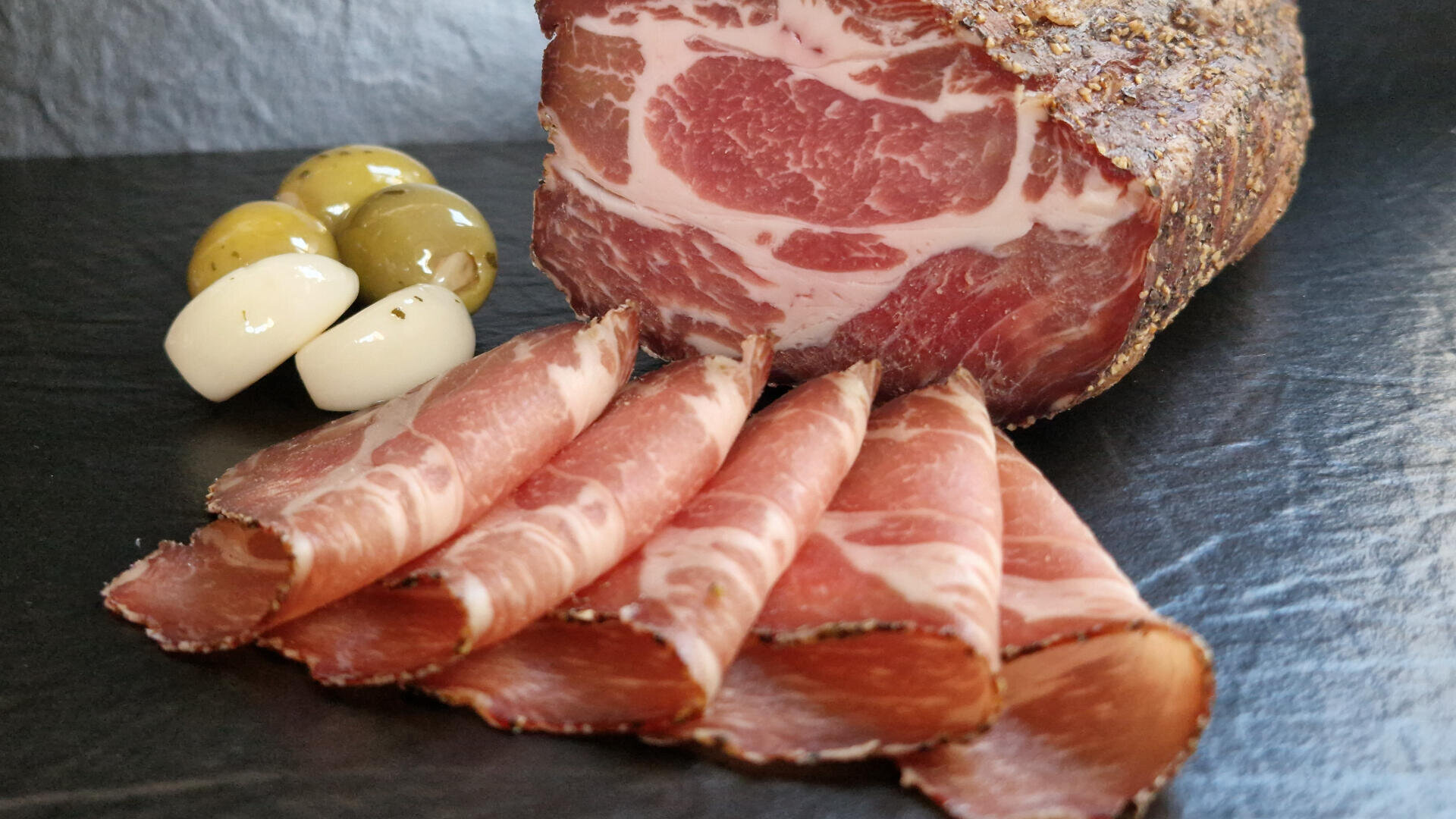 Aufgeschnittener Rohschinken, vor einem größeren Stück angerichtet, dazu grüne und schwarze Oliven sowie zwei Scheiben Weißkäse oder Knoblauch auf einer dunklen Schieferplatte.