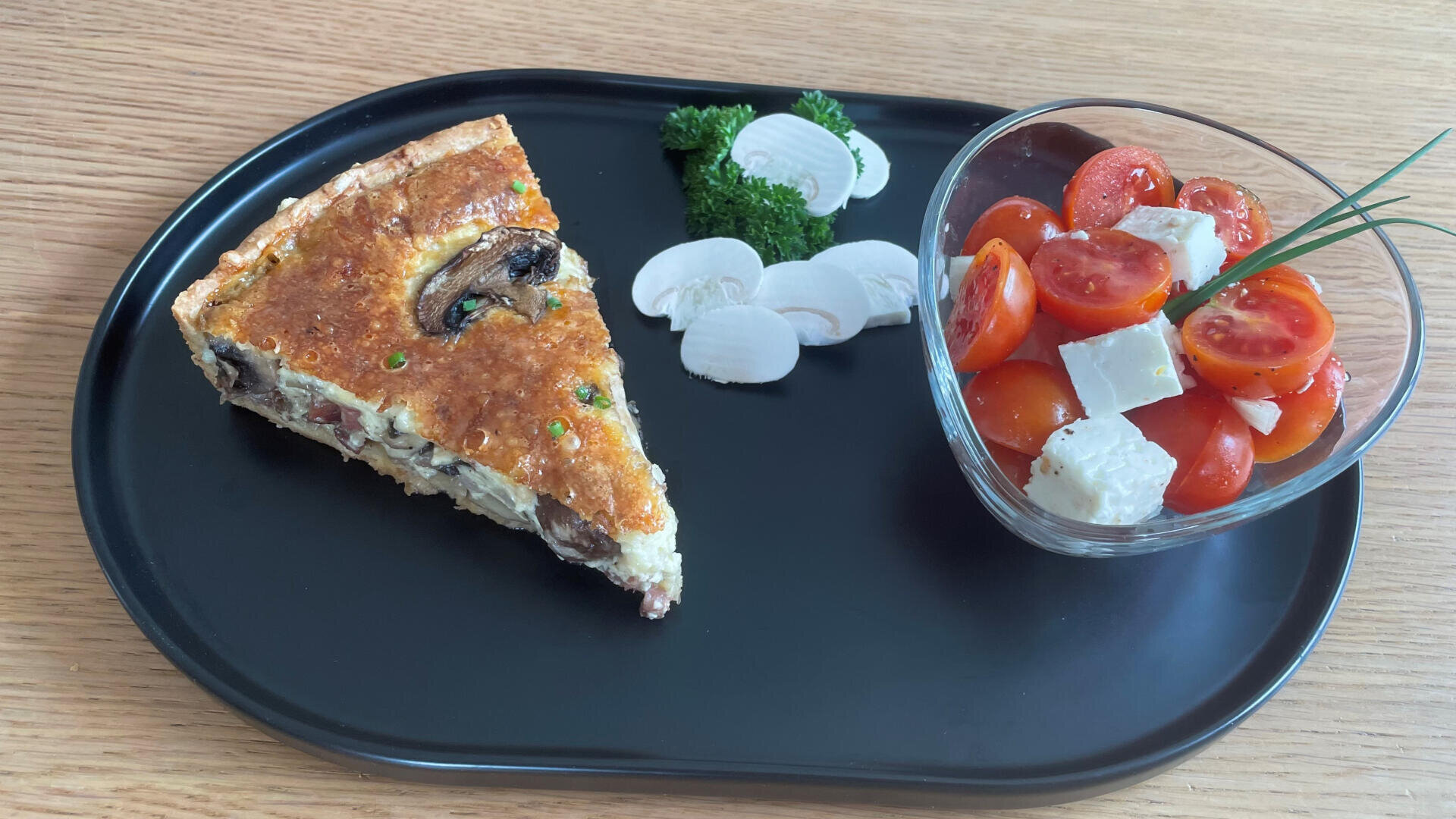 Ein schwarzer Teller mit einer Scheibe Gemüse-Quiche, in Scheiben geschnittenen rohen Pilzen, Petersilie und einer Schale mit Kirschtomaten und Feta-Salat, garniert mit Schnittlauch, auf einem Holztisch.