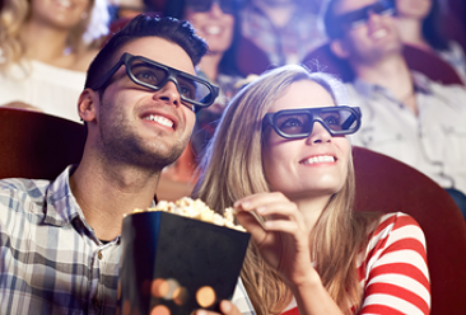 Ein Mann und eine Frau mit 3D-Brille lächeln, während sie sich in einem Kino einen Film ansehen. Die Frau hält eine Schachtel Popcorn in der Hand, und auch die anderen Personen im Hintergrund genießen den Film.