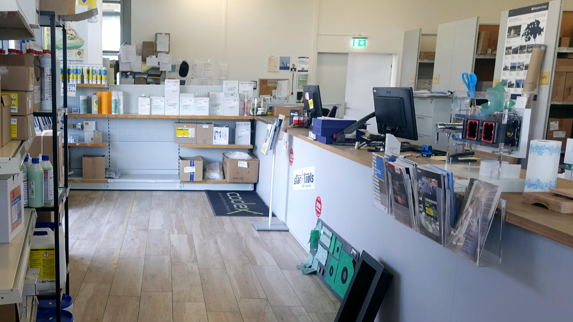 Ein modernes Bürobedarfsgeschäft mit Produktregalen, Computerarbeitsplätzen auf einer Theke, verschiedenen Büromaterialien, Broschüren und Kisten, die ordentlich im Raum verteilt sind.