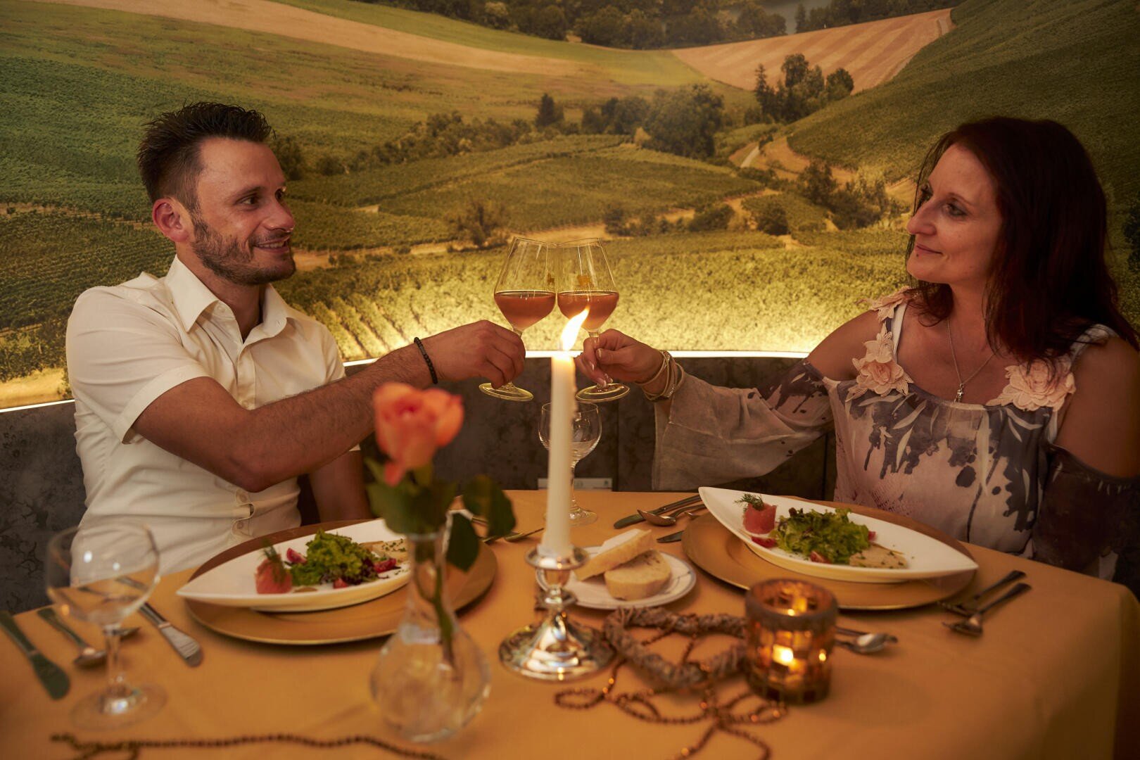 Ein Mann und eine Frau sitzen an einem mit Kerzen beleuchteten Tisch in einem Restaurant und stoßen mit einem Glas Wein an. Sie essen eine Mahlzeit mit Salat, und an der Wand hinter ihnen ist ein ländliches Wandgemälde zu sehen.