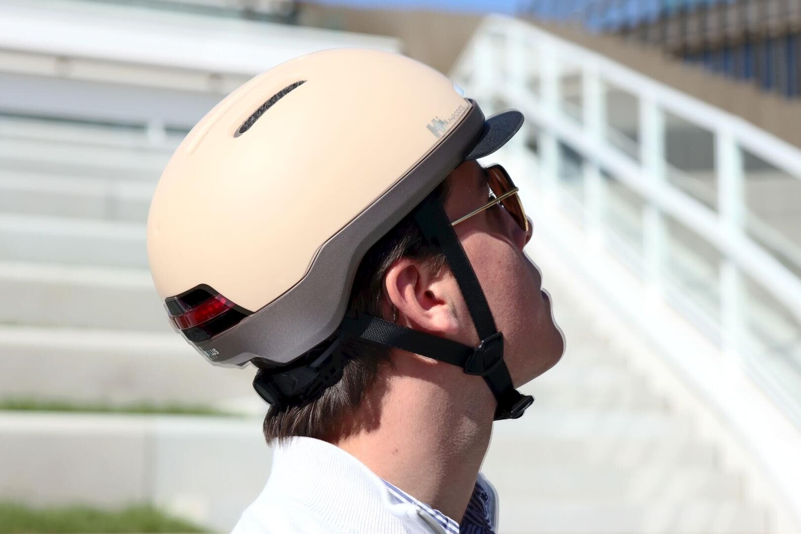 Eine Person, die einen beigen Fahrradhelm und eine Sonnenbrille trägt, blickt im Freien nach oben, mit Treppen und einem weißen Geländer im Hintergrund.