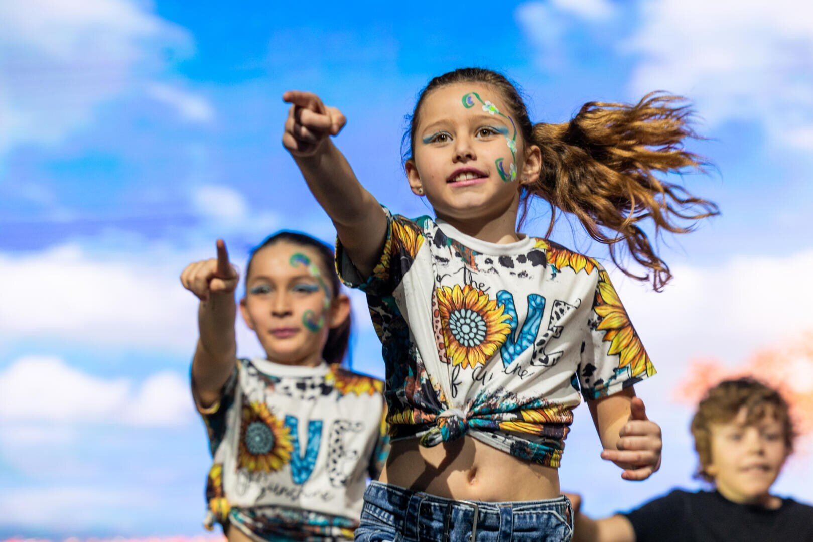 Drei Kinder, die passende Sonnenblumen-T-Shirts und Jeans tragen, tanzen energisch im Freien. Zwei Mädchen im Vordergrund zeigen lächelnd nach vorne, während ein Junge im Hintergrund mitmacht. Der Himmel ist hell mit weichen Wolken hinter ihnen.