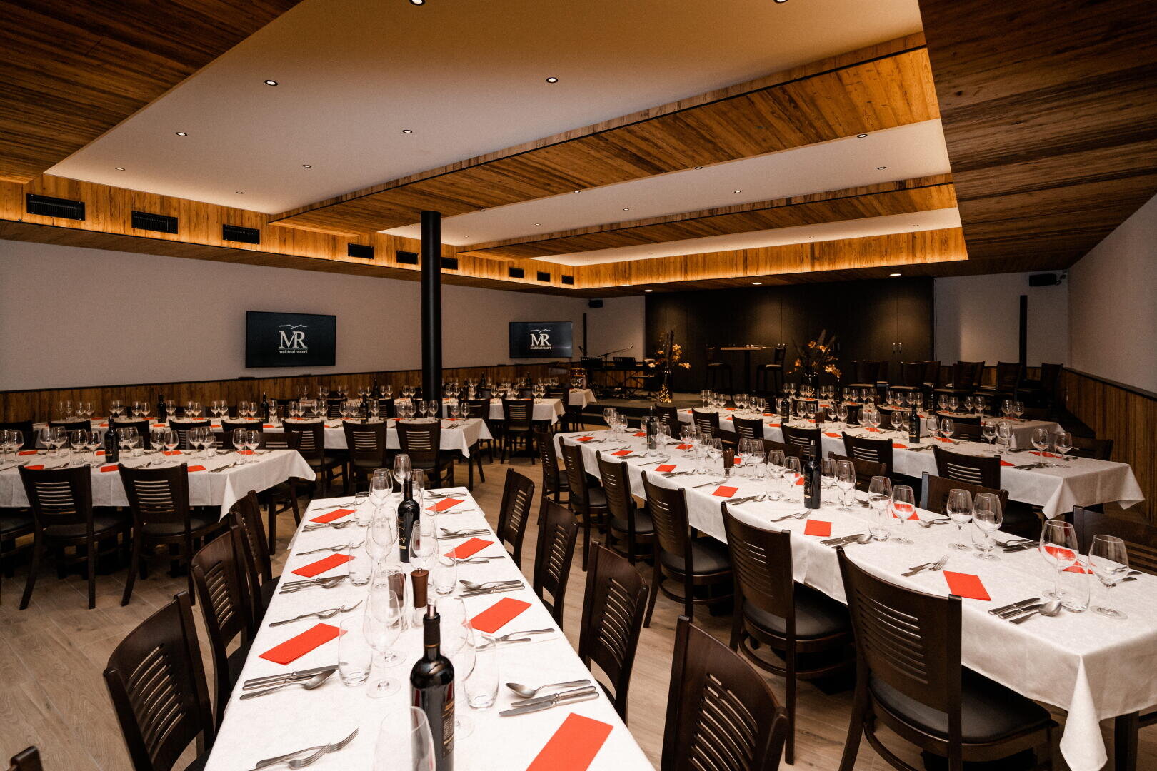 Ein geräumiger, moderner Speisesaal ist für eine Veranstaltung mit langen, weiß gedeckten Tischen, roten Servietten, Weinflaschen, Gläsern und Besteck eingerichtet. Holzdecken und -wände sorgen für eine warme Atmosphäre, und auf Bildschirmen ist das MR-Logo zu sehen.