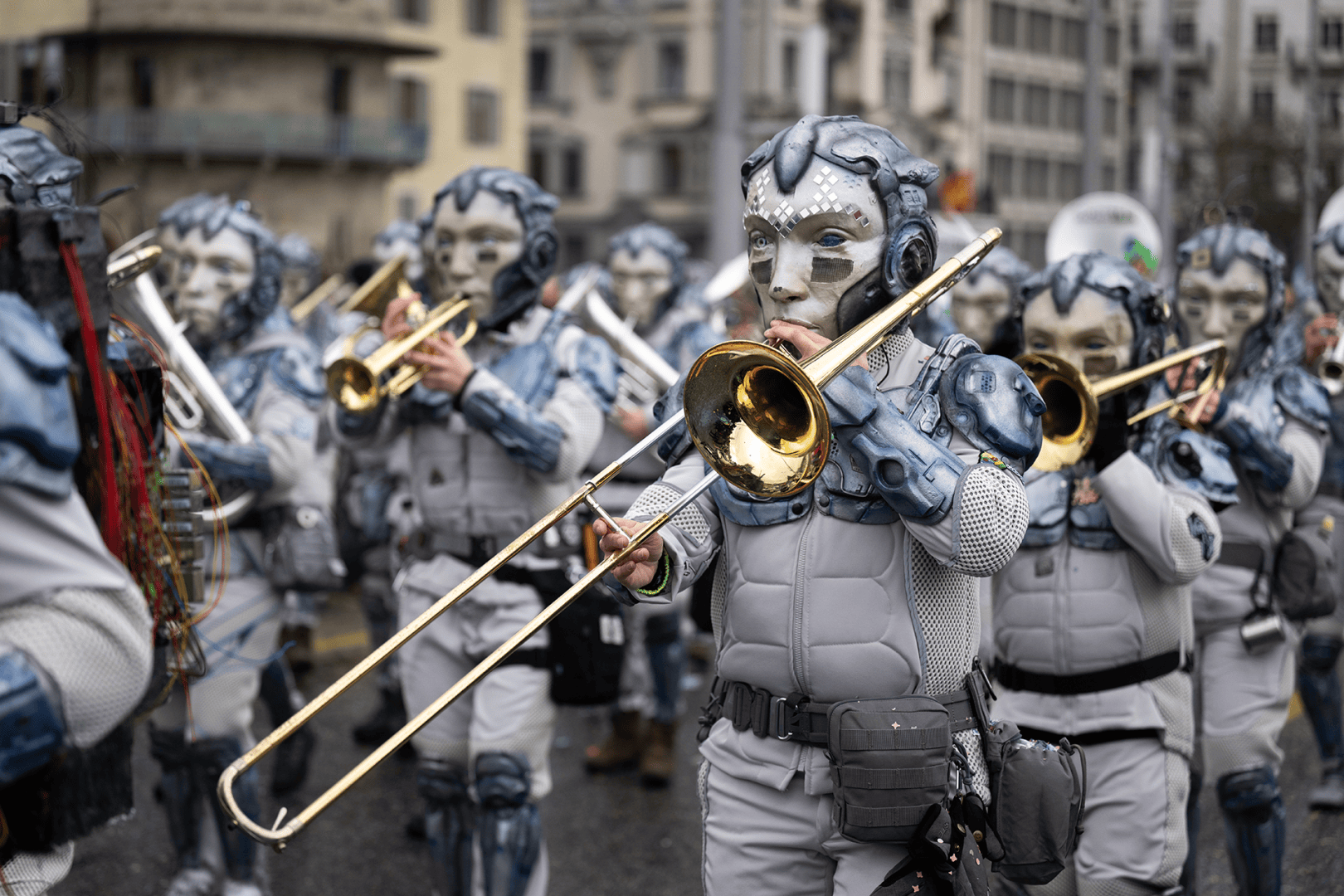 Eine Gruppe von Menschen in futuristischen Roboterkostümen und Masken spielt Blasinstrumente, während sie in einer Stadtparade marschieren. Im Hintergrund sind Gebäude und andere maskierte Darsteller zu sehen.