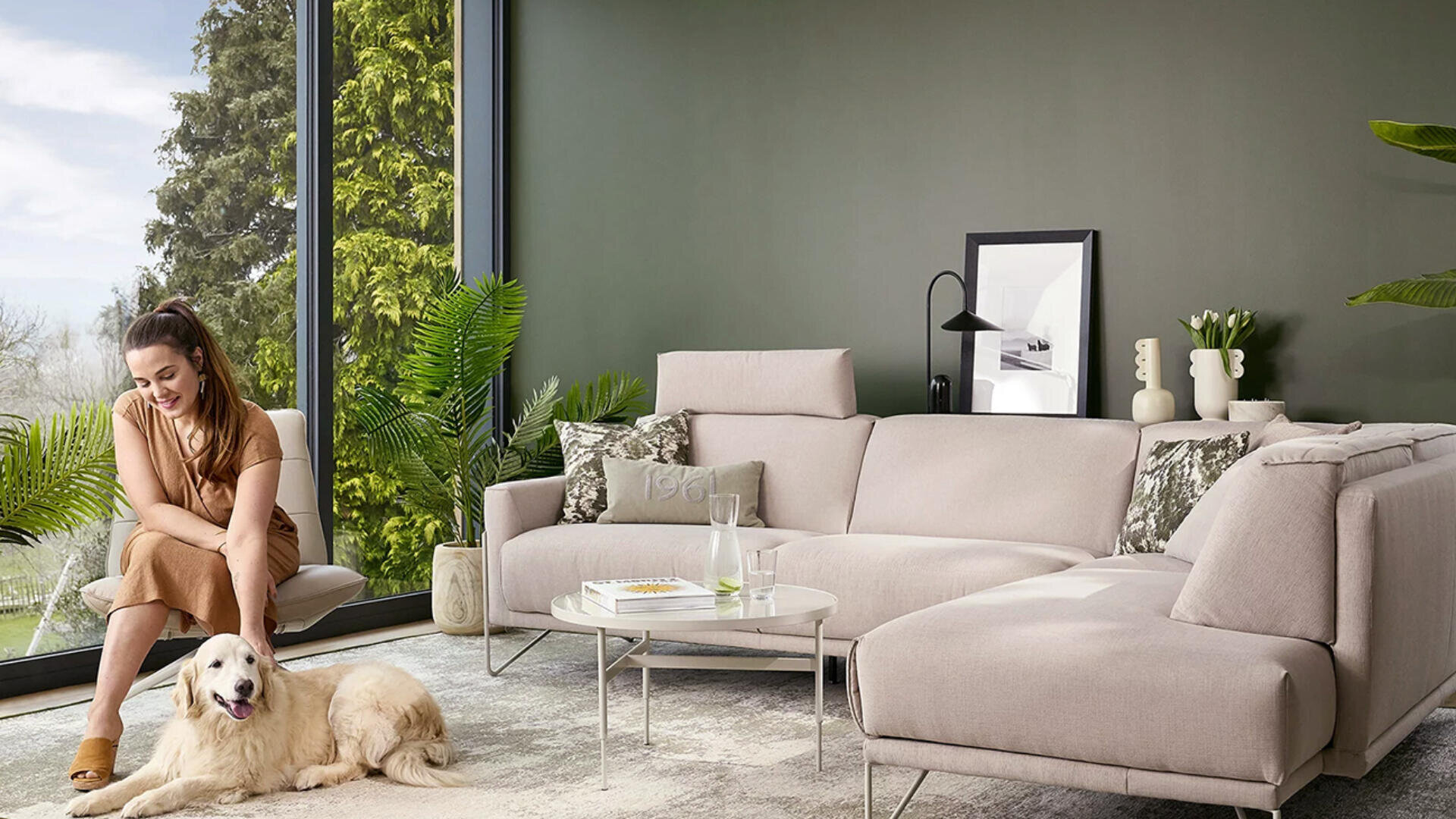 Eine Frau sitzt auf einem beigen Stuhl und streichelt einen Golden Retriever in einem modernen Wohnzimmer mit einem großen Sektionssofa, einem runden Couchtisch und Grünpflanzen an einem hohen Fenster mit Blick auf Bäume.