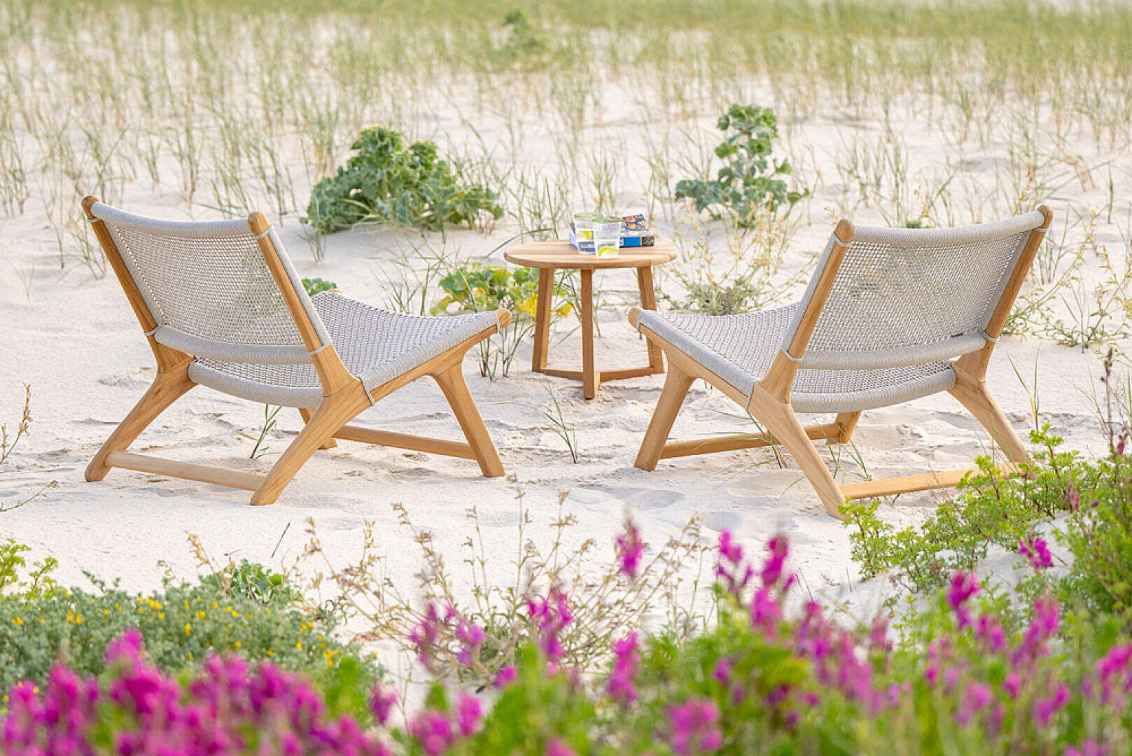 Zwei geflochtene Liegestühle stehen vor einem kleinen runden Holztisch mit Getränken und einem Buch, auf sandigem Boden mit Strandgras und rosa Blumen im Vordergrund.