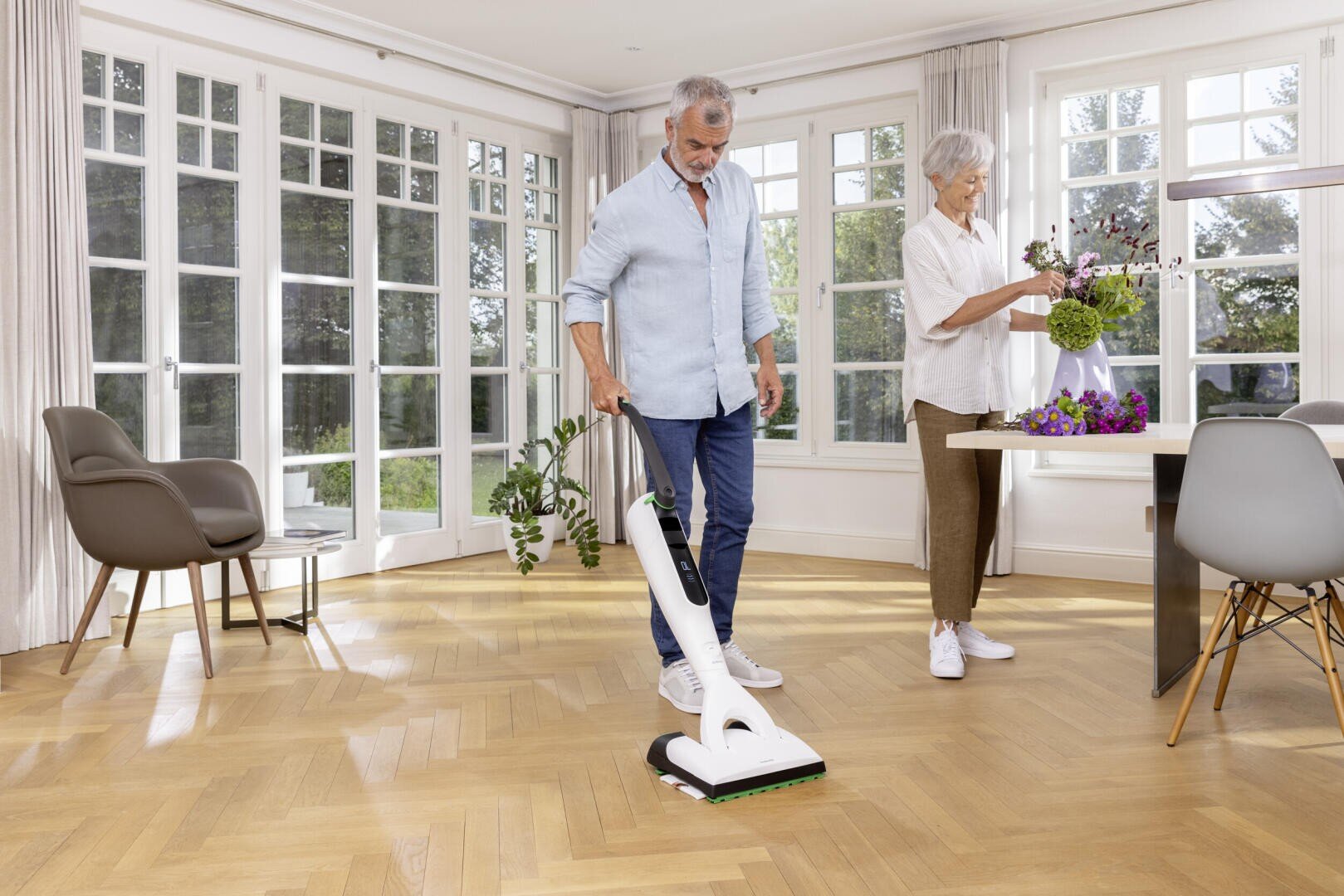 Ein älterer Mann saugt ein helles, modernes Wohnzimmer mit Holzböden, während eine ältere Frau Blumen auf einem Esstisch in der Nähe arrangiert. Große Fenster erfüllen den Raum mit natürlichem Licht.