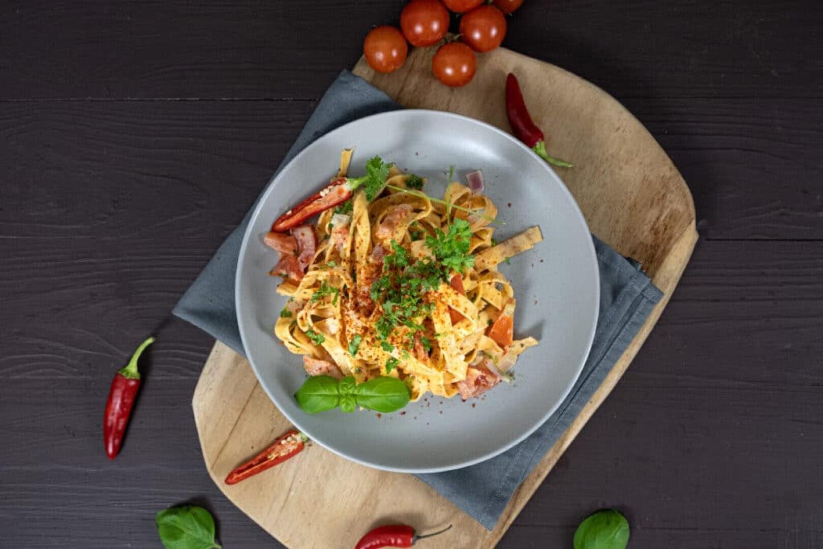 Ein Teller mit Tagliatelle-Nudeln mit Schinken, Tomaten, Chilischoten und frischen Kräutern steht auf einer grauen Serviette auf einem Holzbrett, umgeben von Kirschtomaten, roten Chilischoten und Basilikumblättern.
