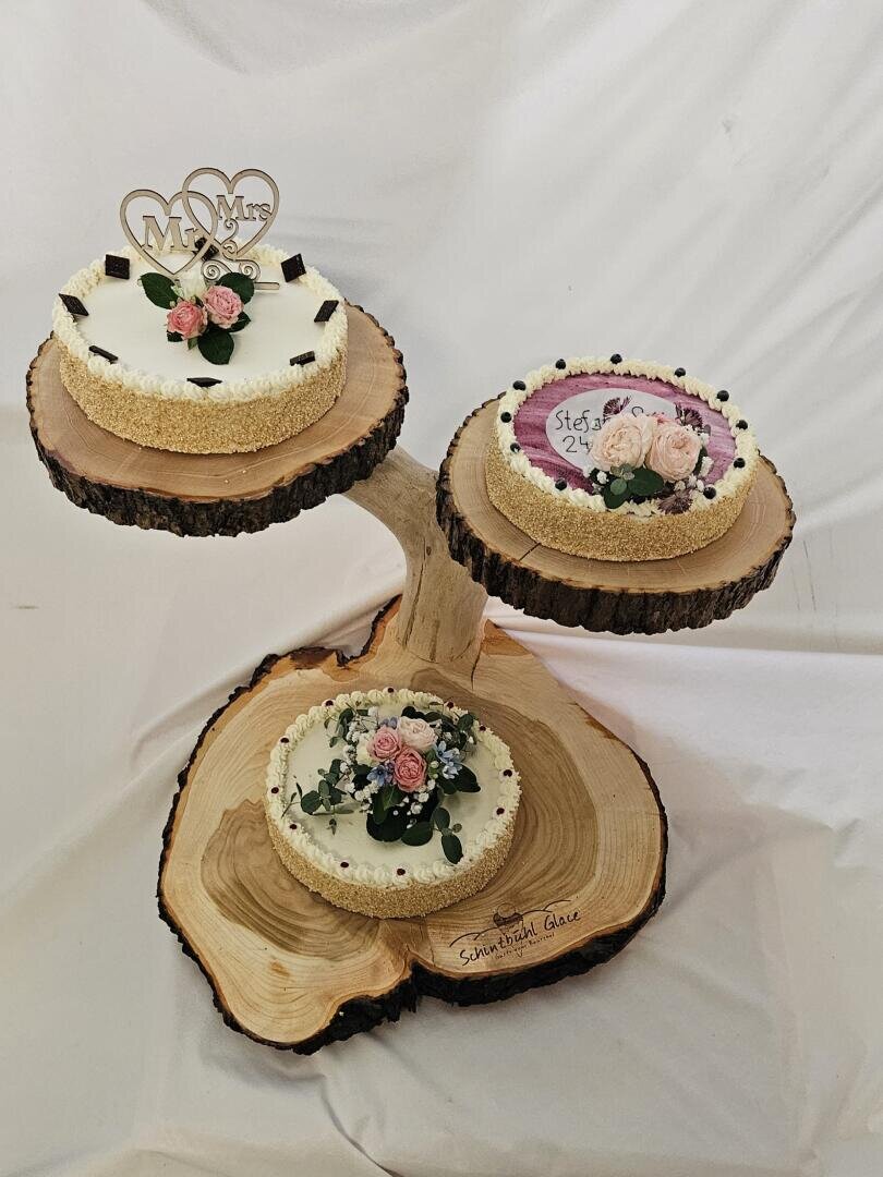 Drei runde Torten auf einem rustikalen Holzkuchenständer. Die Torten sind mit Blumen verziert, und eine hat einen Mr & Mrs Topper. Ein anderer Kuchen hat lila Zuckerguss mit Text und Blumendekoration. Weißer Hintergrund.