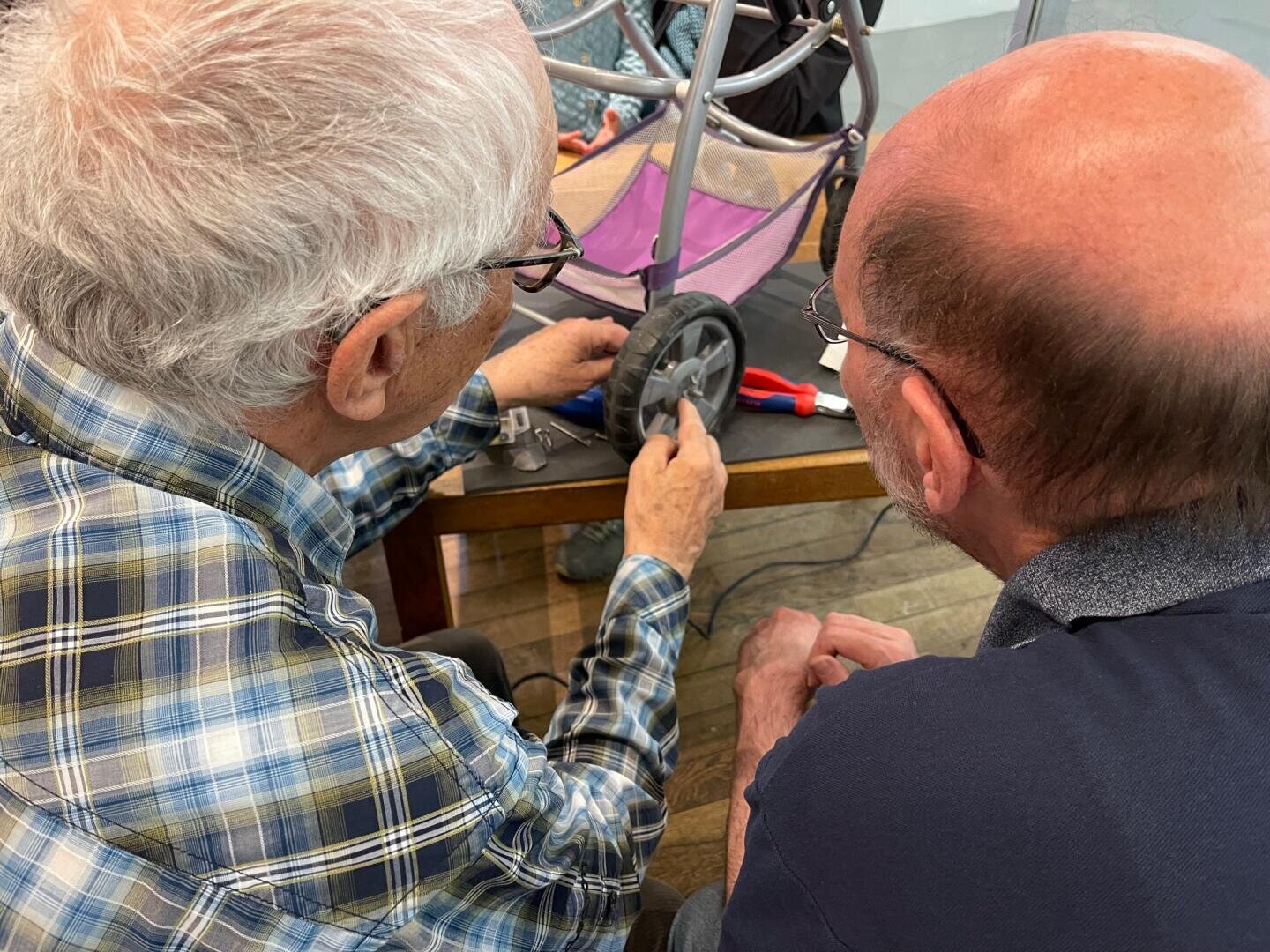 Zwei ältere Männer sitzen an einem Tisch und arbeiten gemeinsam an der Reparatur eines kleinen Rades. Ein Mann hält und untersucht das Rad, während der andere es genau beobachtet. Auf dem Tisch in der Nähe sind Werkzeuge zu sehen.