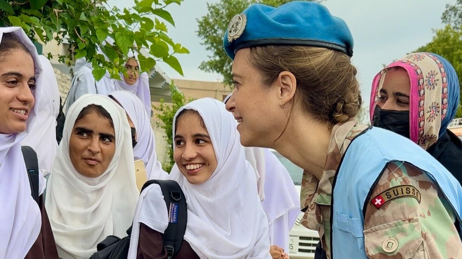 Ein lächelnder UN-Friedenswächter in Uniform unterhält sich im Freien mit einer Gruppe von Mädchen in weißen Kopftüchern und braunen Uniformen. Die Mädchen lächeln und sind beschäftigt, und im Hintergrund sind Bäume und Gebäude zu sehen.