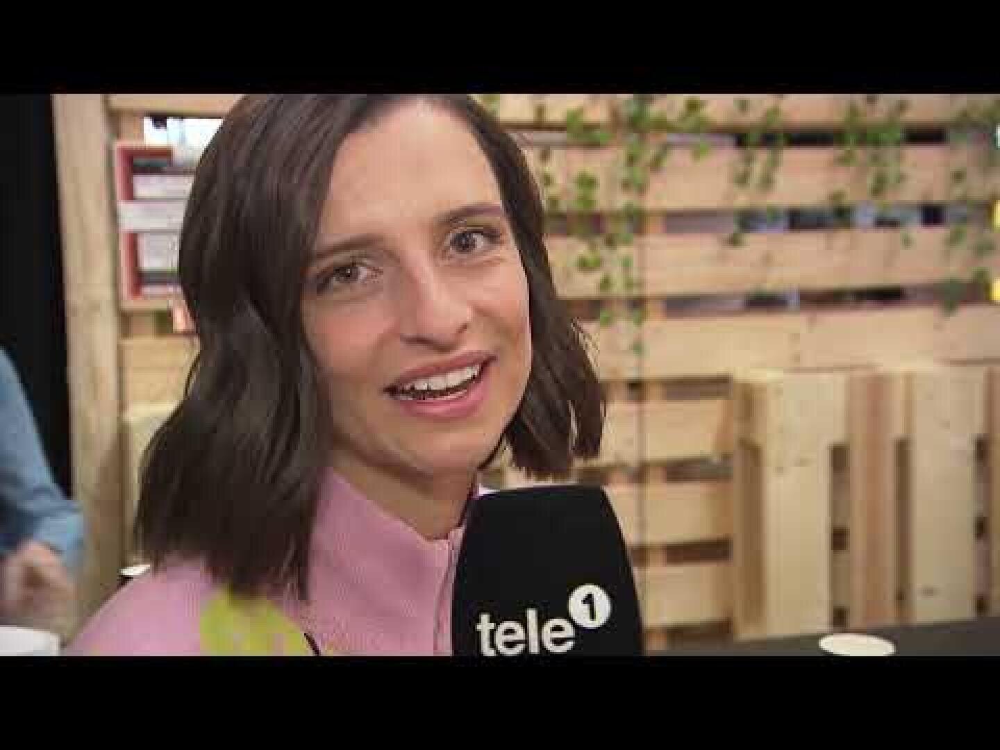 Eine Frau mit schulterlangen braunen Haaren lächelt in die Kamera und hält ein Mikrofon mit der Aufschrift tele 1. Sie steht in einem Innenraum vor einer Kulisse aus Holzpaletten, die mit grünen Pflanzen geschmückt ist.