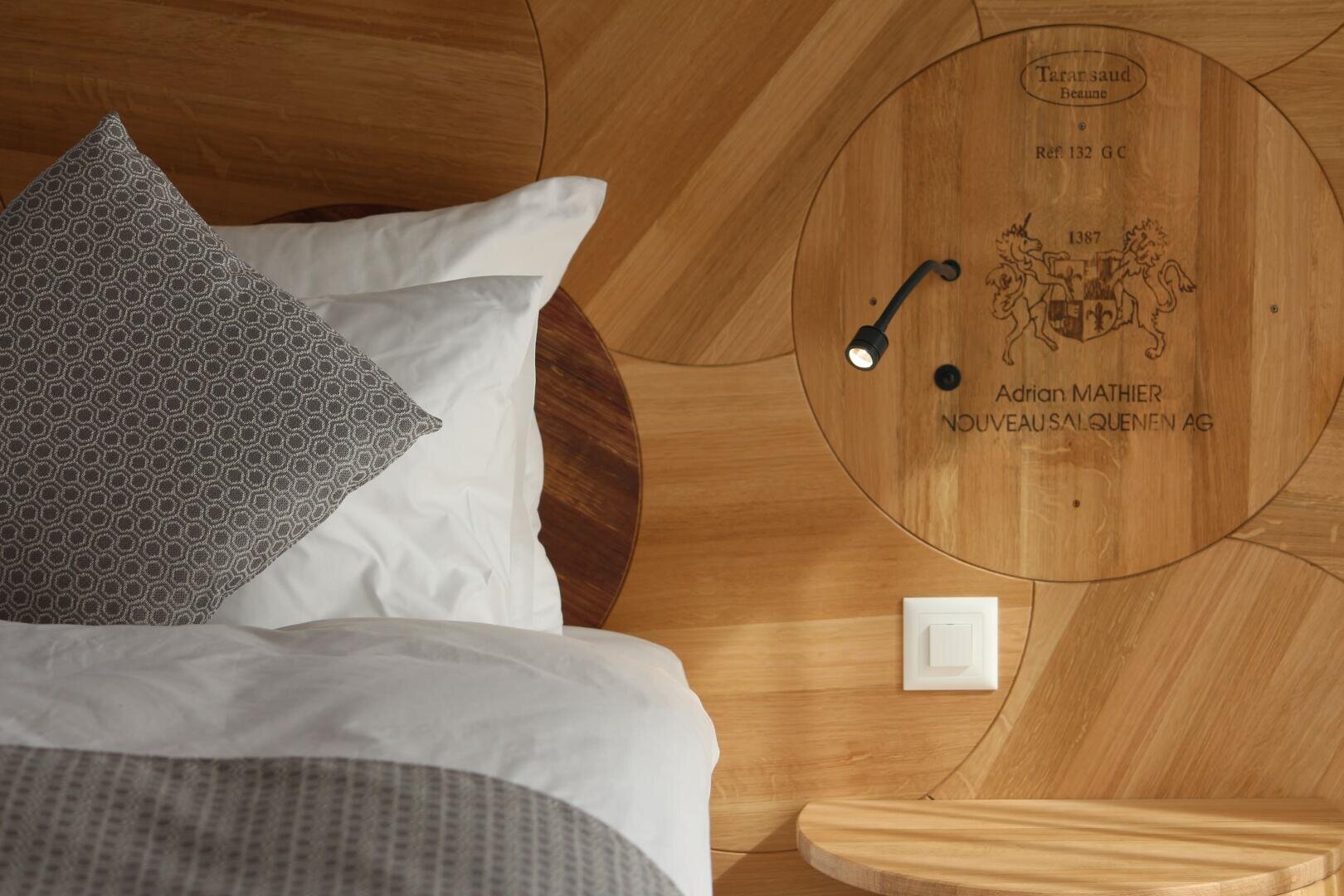 Ein modernes, gemütliches Bett mit weißer Bettwäsche und gemusterten Kissen neben einer Holzwand mit einem kreisförmigen, eingravierten Muster, einer kleinen Leselampe und einem Lichtschalter.
