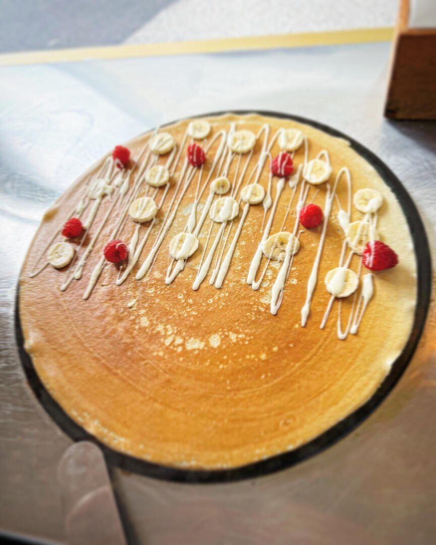 Ein großer, dünner Crêpe auf einem runden Grill, belegt mit Bananenscheiben, Himbeeren und mit weißer, gestreifter Soße beträufelt. Der Hintergrund ist leicht unscharf.