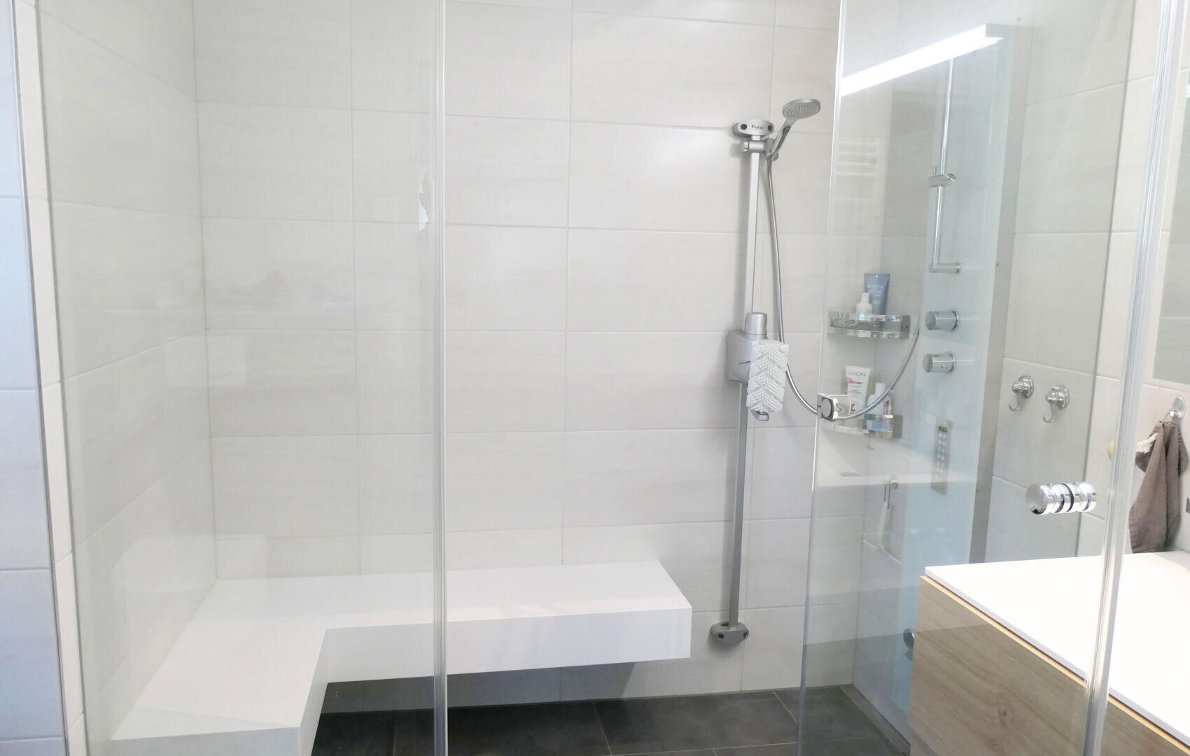 Moderne, begehbare Dusche mit eingebauter weißer Sitzbank, Handbrause, Glastüren und Ablageflächen für Toilettenartikel; die Wände sind mit beigen und der Boden mit dunklen Fliesen belegt.
