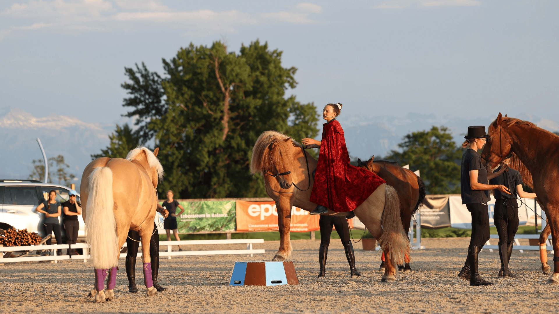 Eine Person mit rotem Umhang und Krone sitzt auf einem braunen Pferd in einer Freiluftarena, umgeben von drei anderen Pferden und Pferdeführern. Im Hintergrund sind Bäume und Berge zu sehen.
