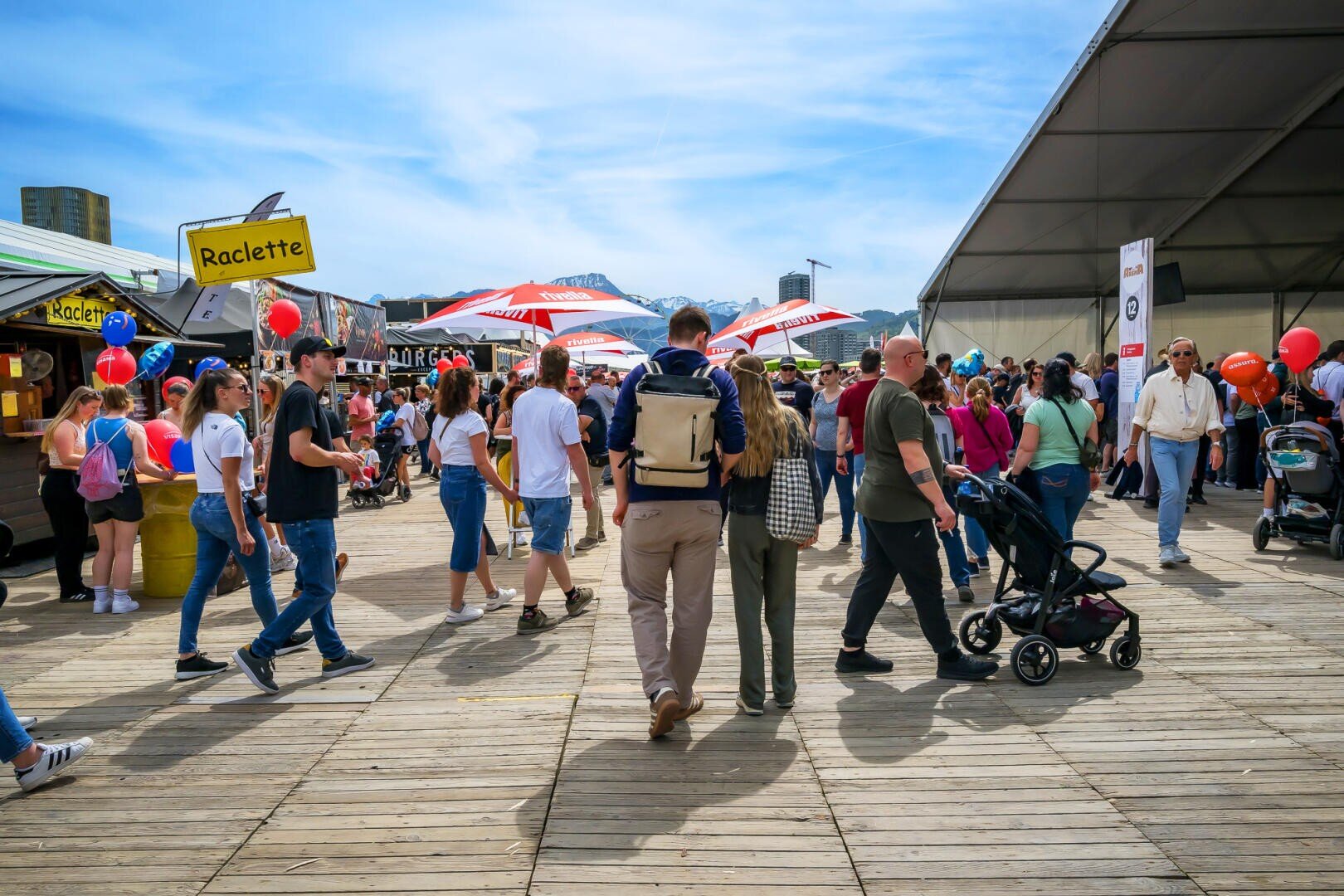 Eine lebhafte Festivalkulisse mit Menschen, die auf Holzbrettern laufen, Essensständen mit der Aufschrift "Raclette", Regenschirmen, Spaziergängern und Luftballons. Im Hintergrund sind Berge und blauer Himmel zu sehen.