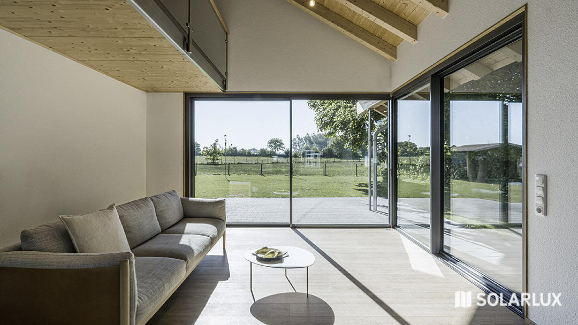 Modernes Wohnzimmer mit großen, raumhohen Glasschiebetüren, einem grauen Sofa, einem kleinen weißen Couchtisch und einer Holzdecke. Der Raum öffnet sich zu einer sonnigen Terrasse und einem ausgedehnten grünen Rasen draußen. Das SOLARLUX-Logo ist sichtbar.
