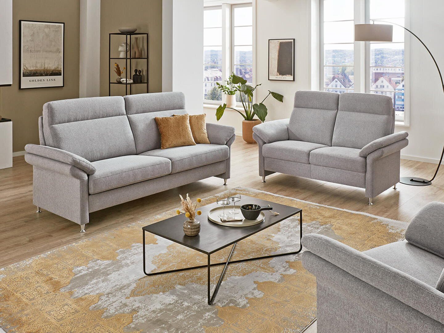 Helles Wohnzimmer mit zwei hellgrauen Sofas, einem modernen schwarzen Couchtisch, einem beige gemusterten Teppich, Topfpflanzen, gerahmter Kunst an den Wänden, großen Fenstern und einer geschwungenen Stehlampe.
